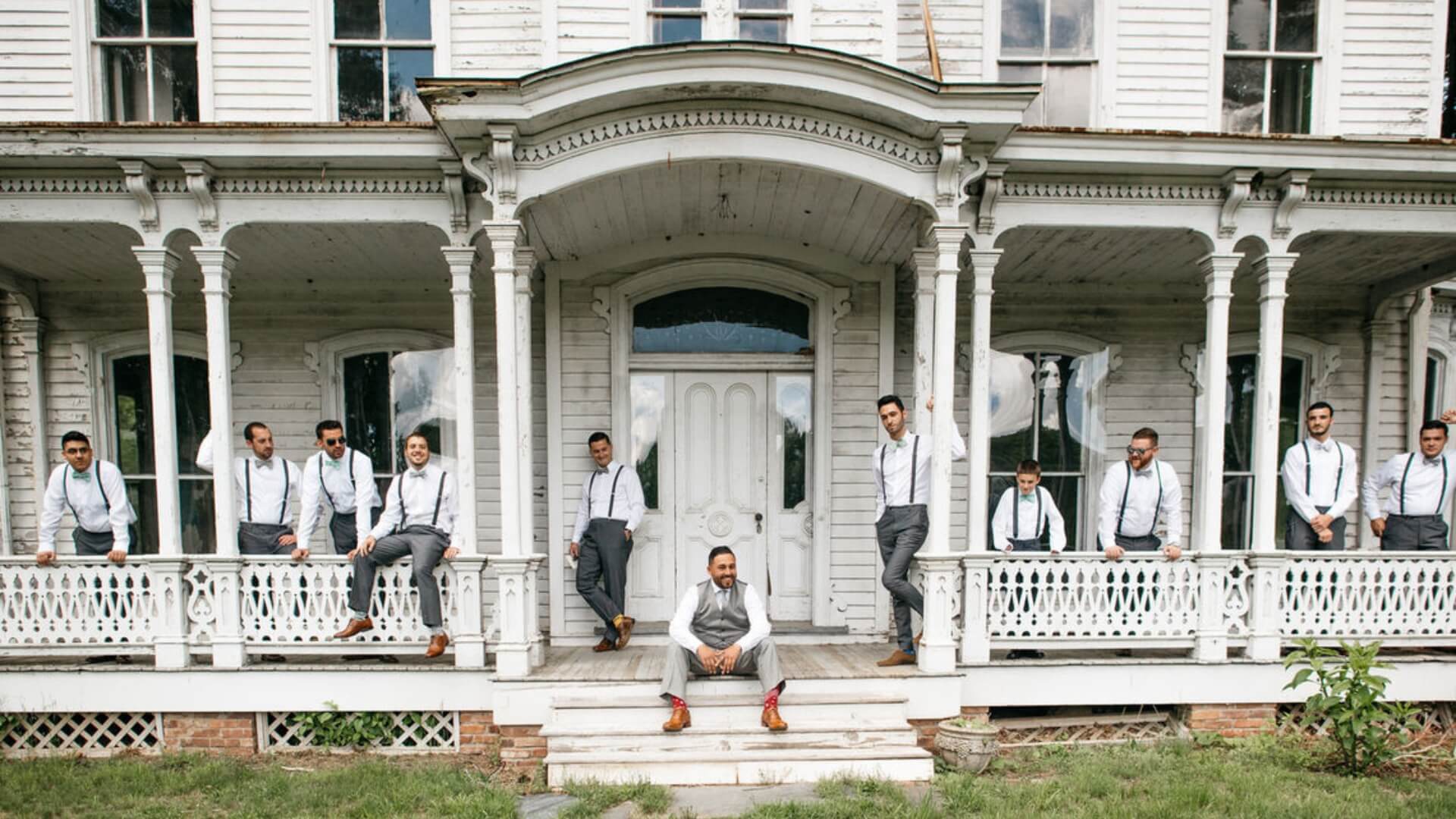A group of men in formal attire with suspenders pose on the porch of a large, old, white wooden house.