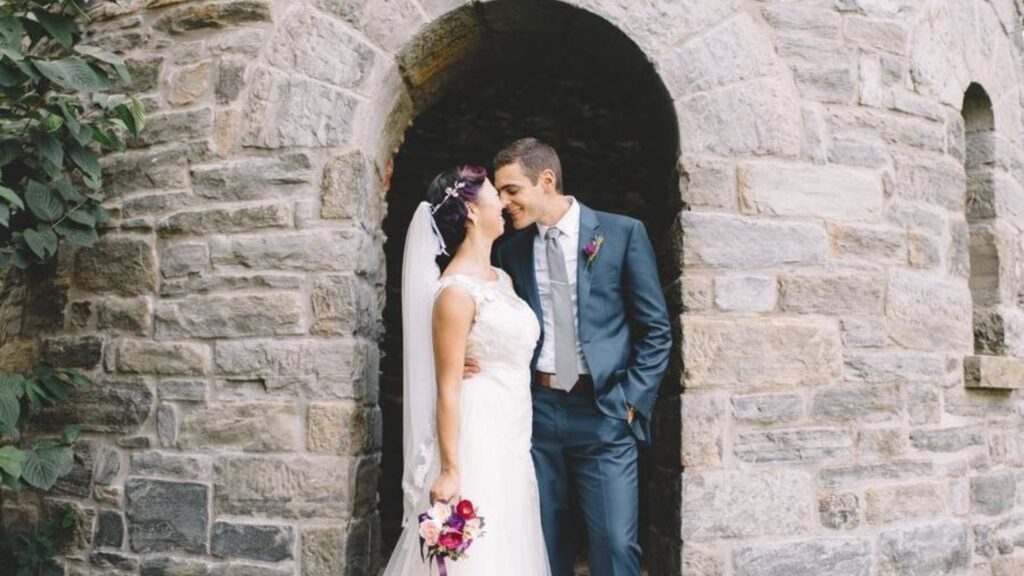 Bride and groom smiling at each other under a stone archway. Bride holds a bouquet, and groom wears a blue suit.