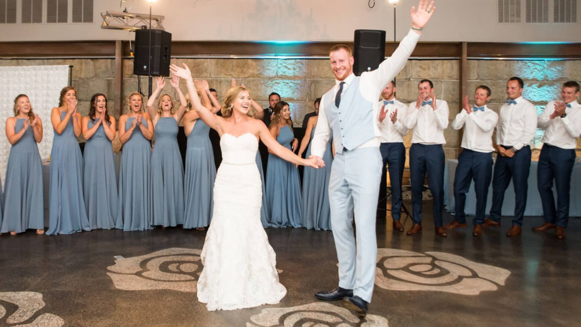 A bride and groom raise their arms while dancing, surrounded by a clapping bridal party in blue dresses and suits.