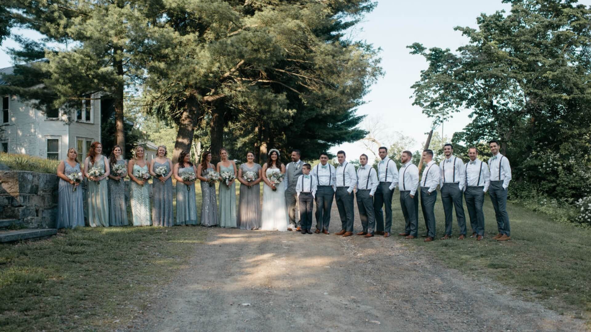 A large wedding party stands in a line outdoors on a dirt path, surrounded by trees. Bridesmaids wear matching dresses, and groomsmen wear shirts with suspenders.
