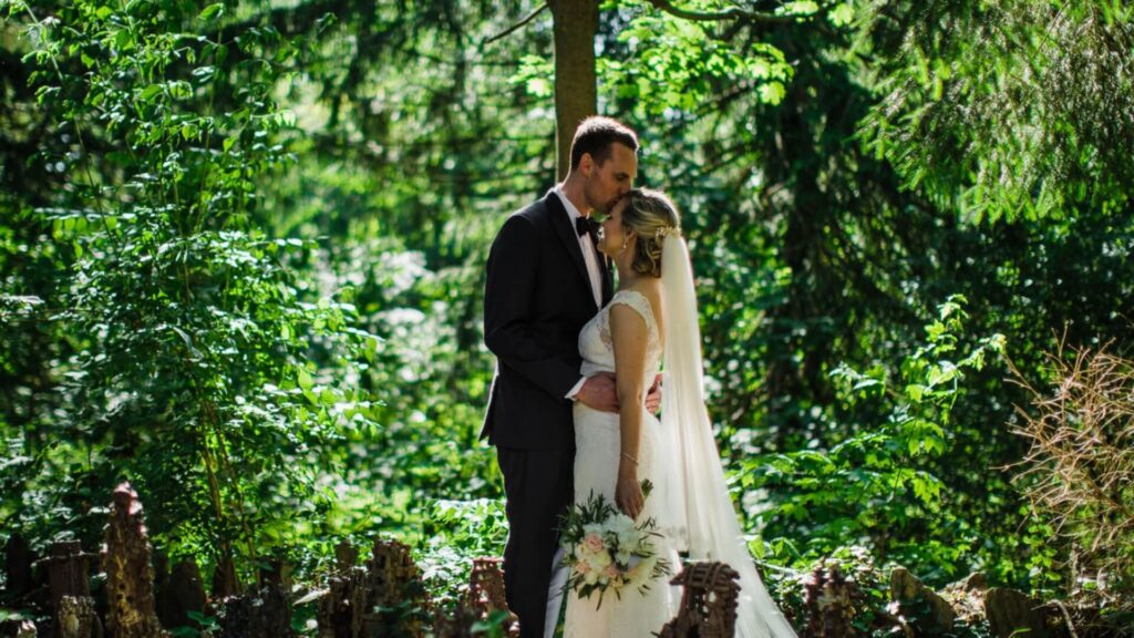 Bride and groom stand closely in a lush green forest setting. The bride holds a bouquet and wears a long veil.