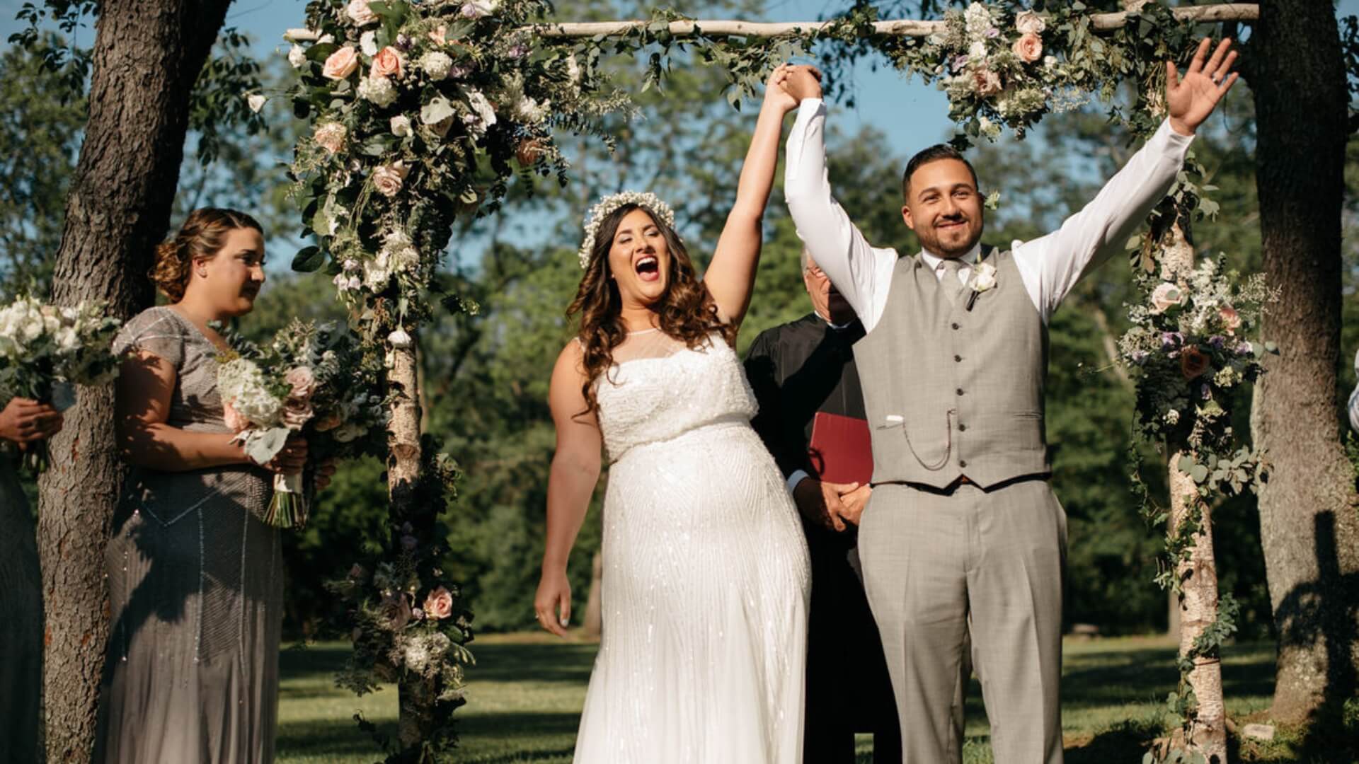 A bride and groom celebrate under a floral arch, raising their hands in joy, with a bridesmaid holding flowers nearby in an outdoor setting.