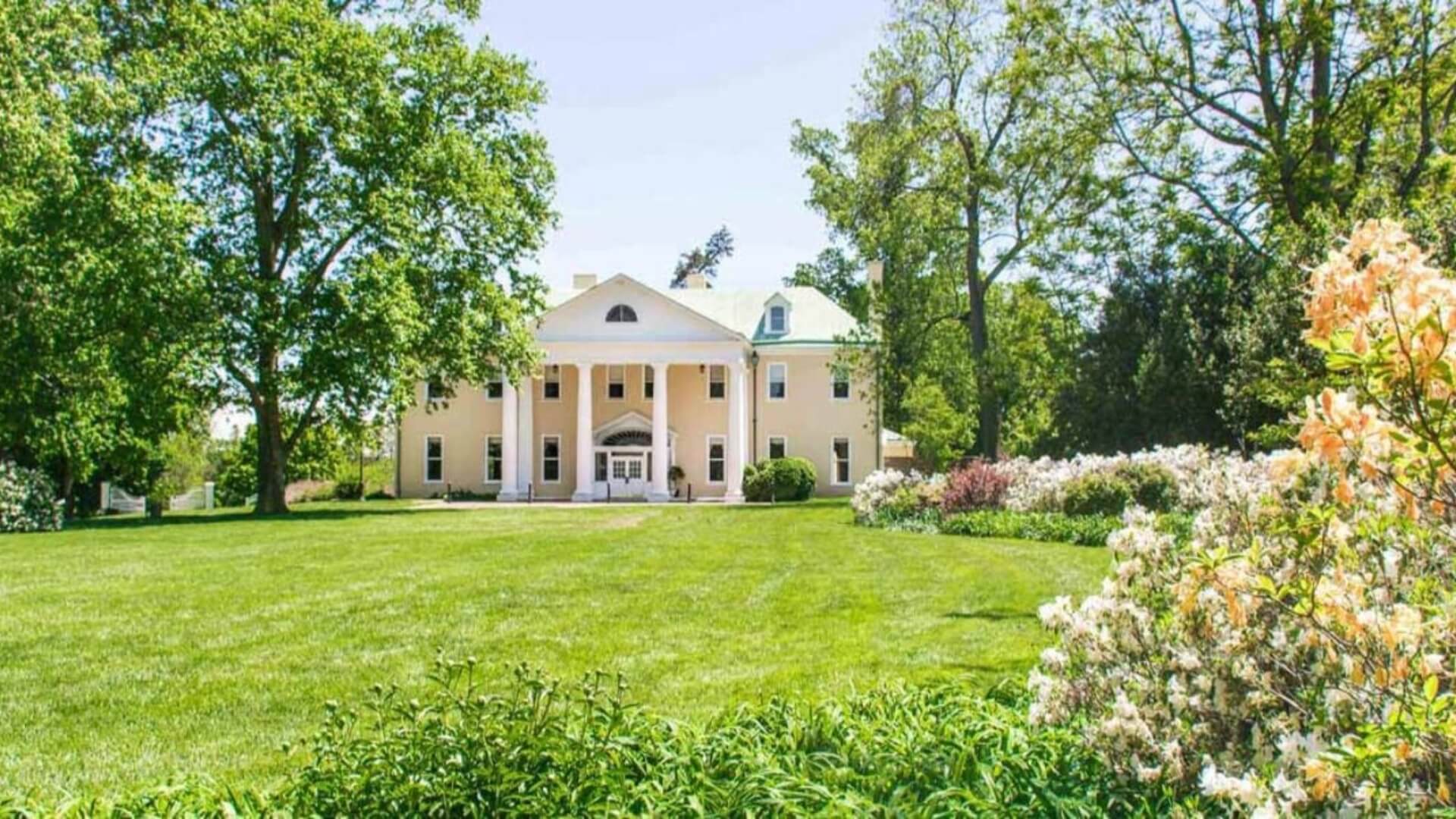 Large yellow mansion with white columns, surrounded by lush green lawn and trees, under a clear blue sky.