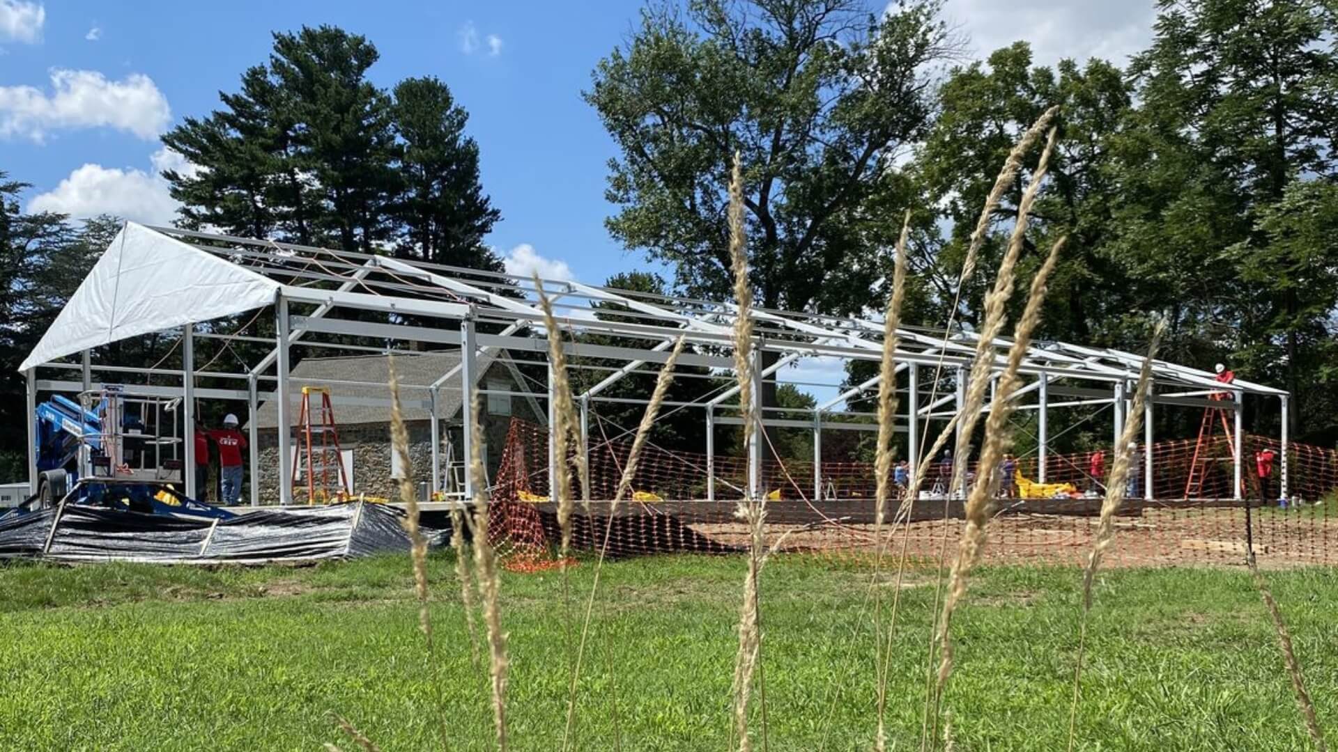 Construction site with metal framework for a building under blue sky, surrounded by trees. Workers and equipment are visible. Tall grass is in the foreground.