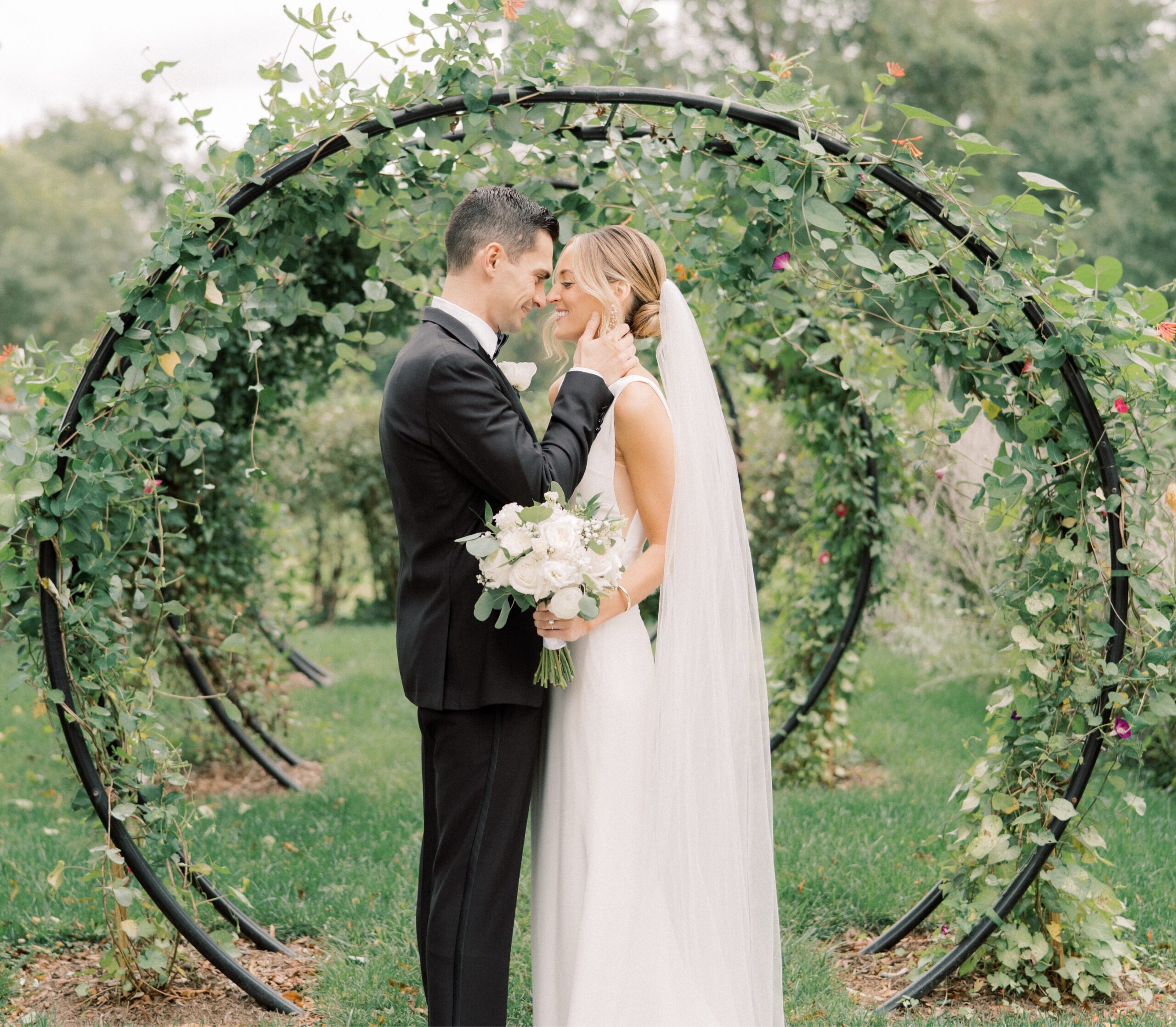A bride and groom stand under a circular, vine-covered archway in a garden. The bride holds a bouquet, and both are dressed in formal wedding attire.