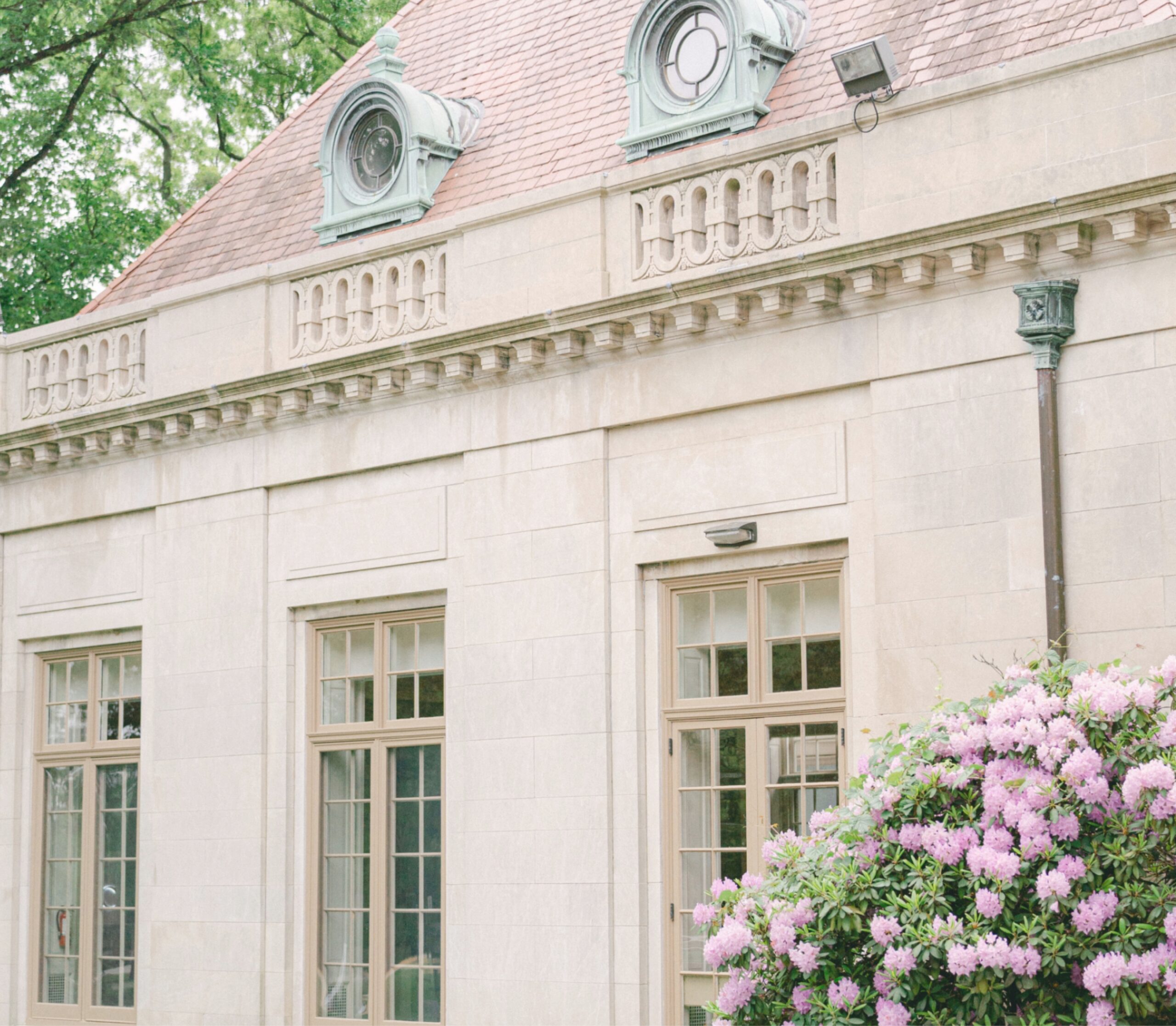 A stone building with round attic windows and ornate trim. Large rectangular windows are below. A blooming bush with pink flowers is in the foreground.