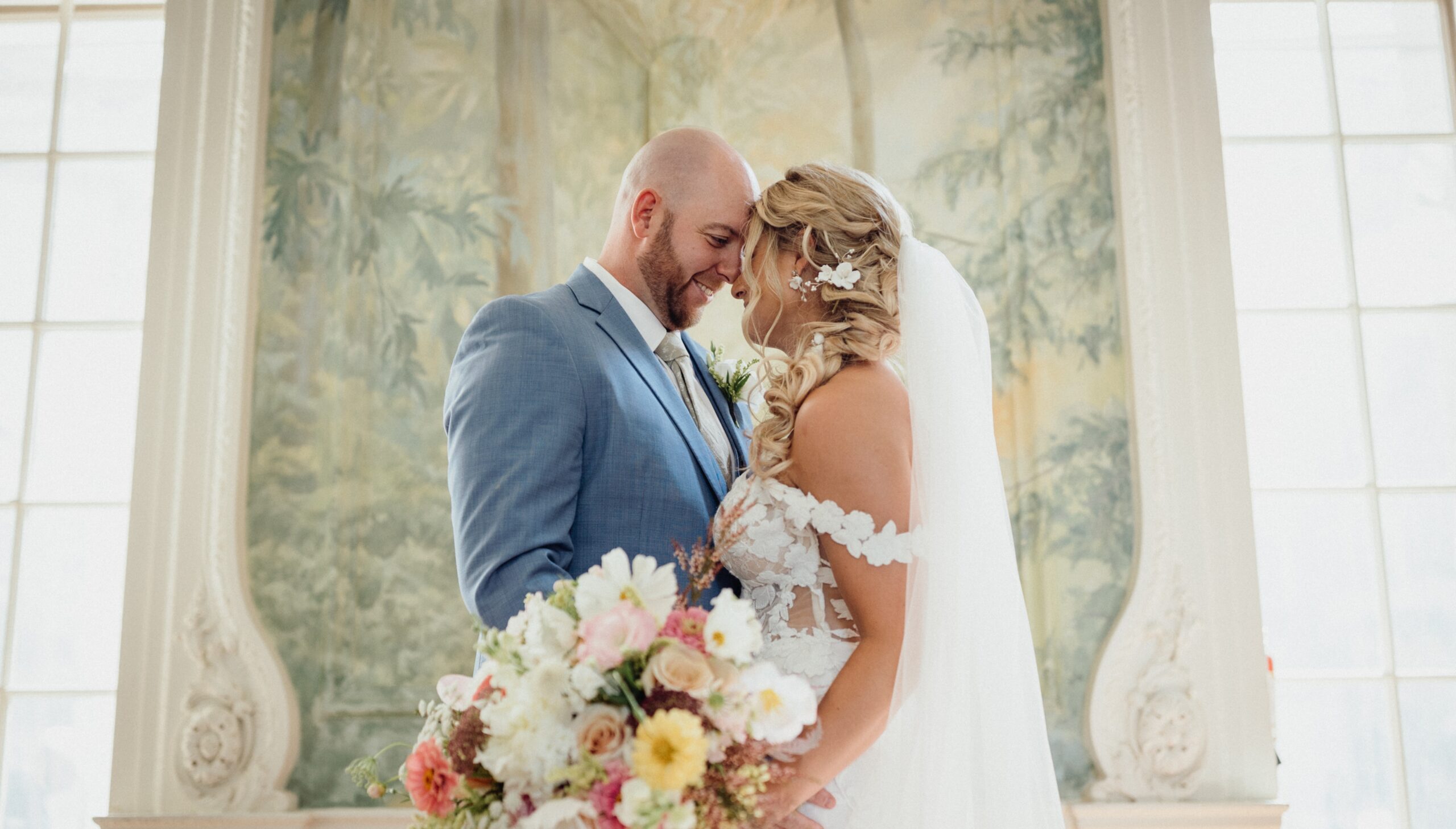 A bride and groom stand closely, smiling. The bride holds a bouquet of flowers. They are indoors with a mural and large windows in the background.