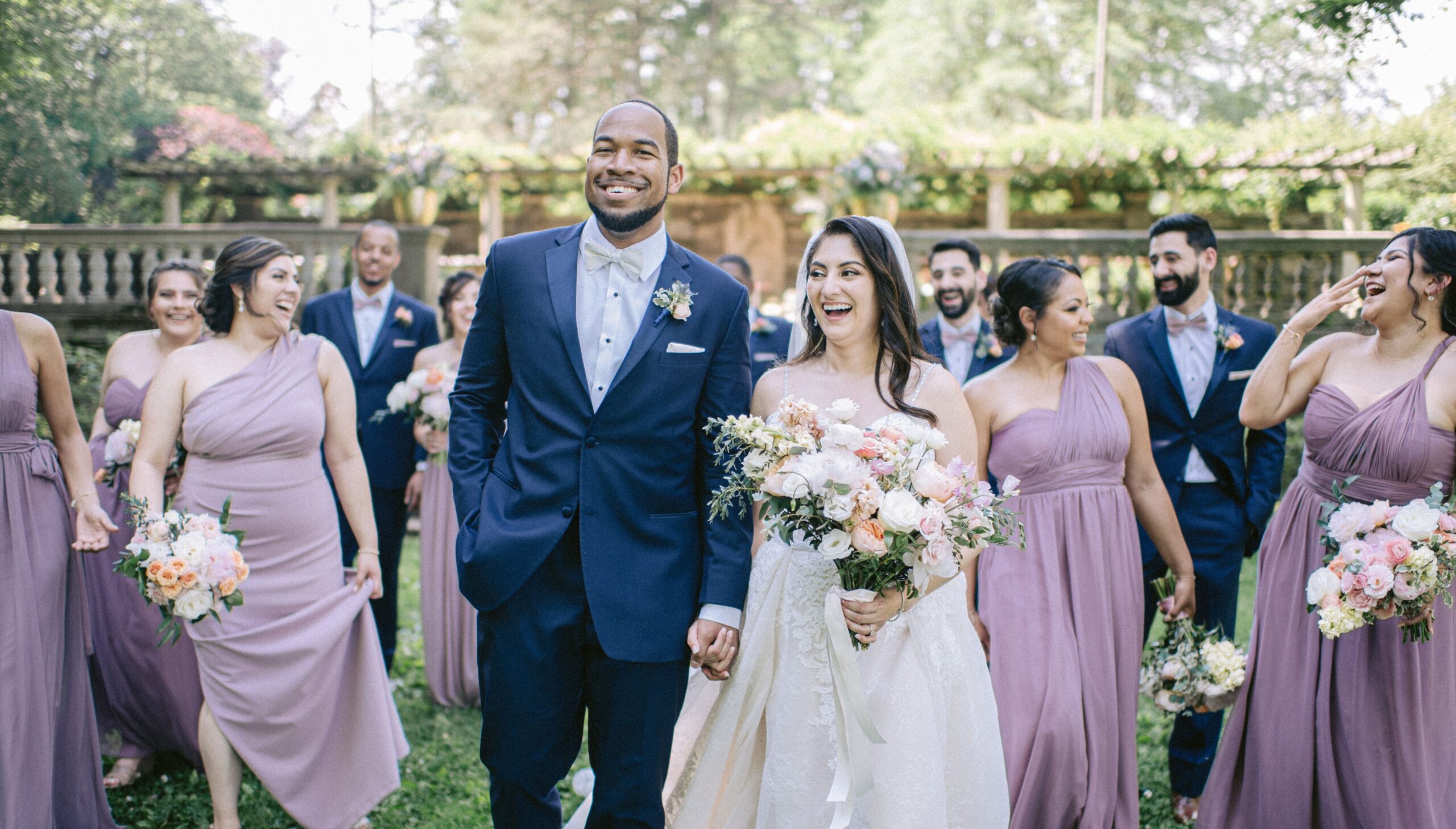 A bride and groom walk outdoors, smiling, with a bridal party in lavender attire surrounding them. They hold flower bouquets.