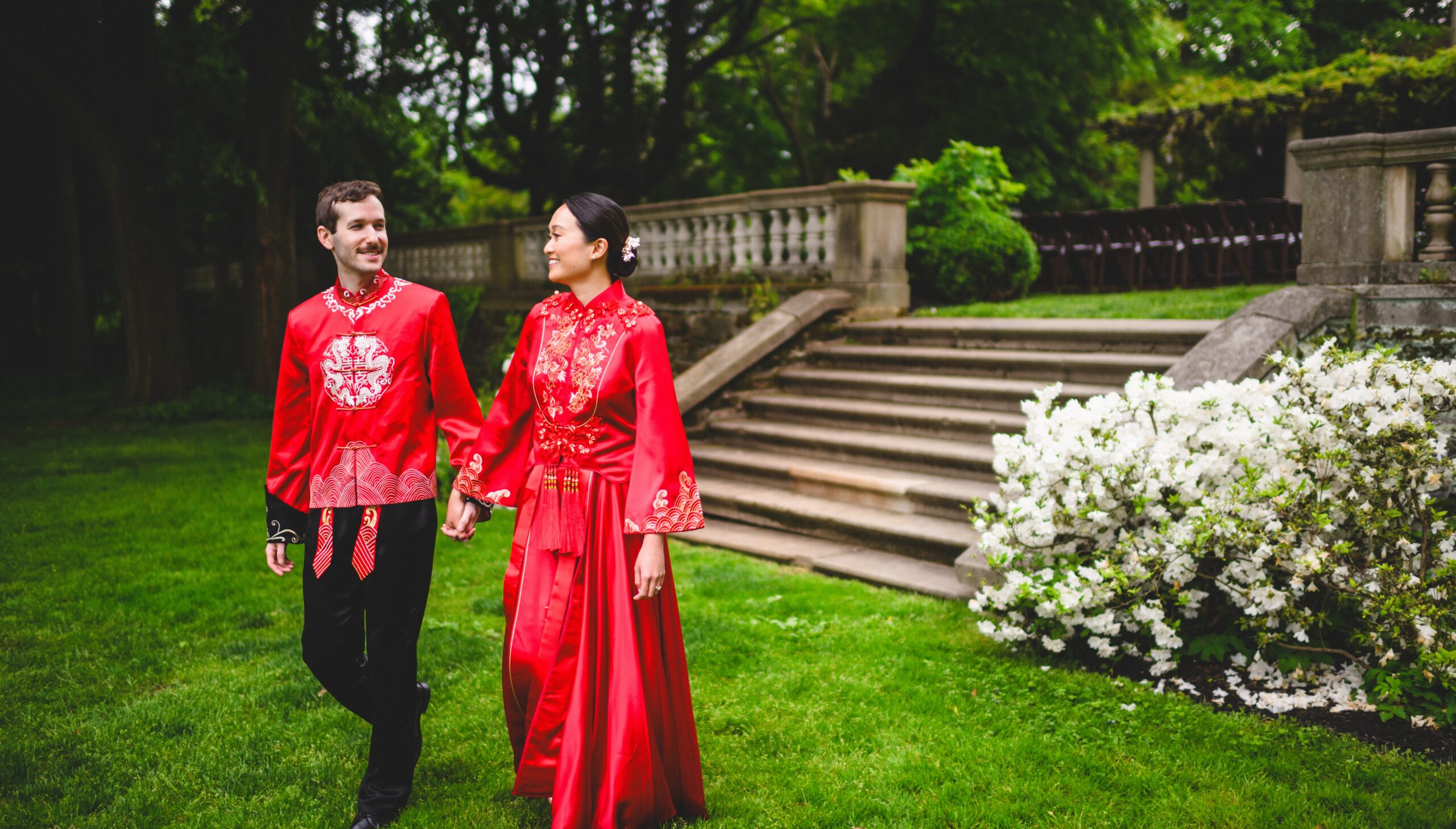 A couple in red traditional attire walks hand in hand on a lawn, with a staircase and white flowers in the background.