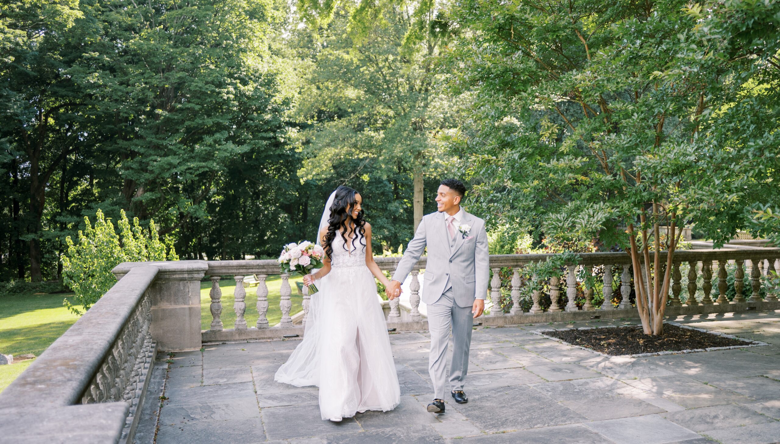 Bride and groom walking hand in hand on a stone terrace, surrounded by lush greenery and trees. The bride holds a bouquet, and both are smiling at each other.