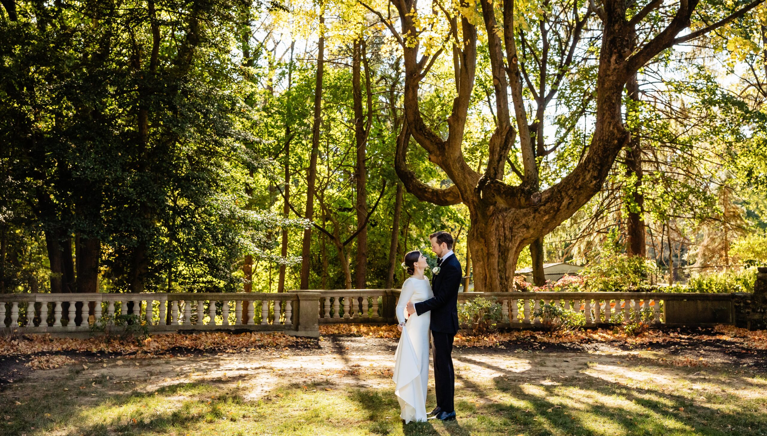 A couple in wedding attire stands facing each other in a sunlit park with tall trees and a stone balustrade in the background.