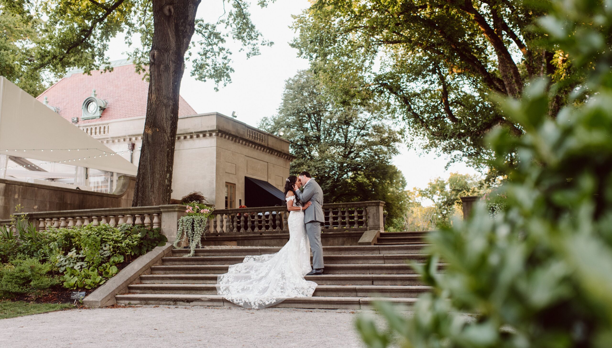 A couple in wedding attire embraces on stone steps in a garden setting, with trees and a building in the background.
