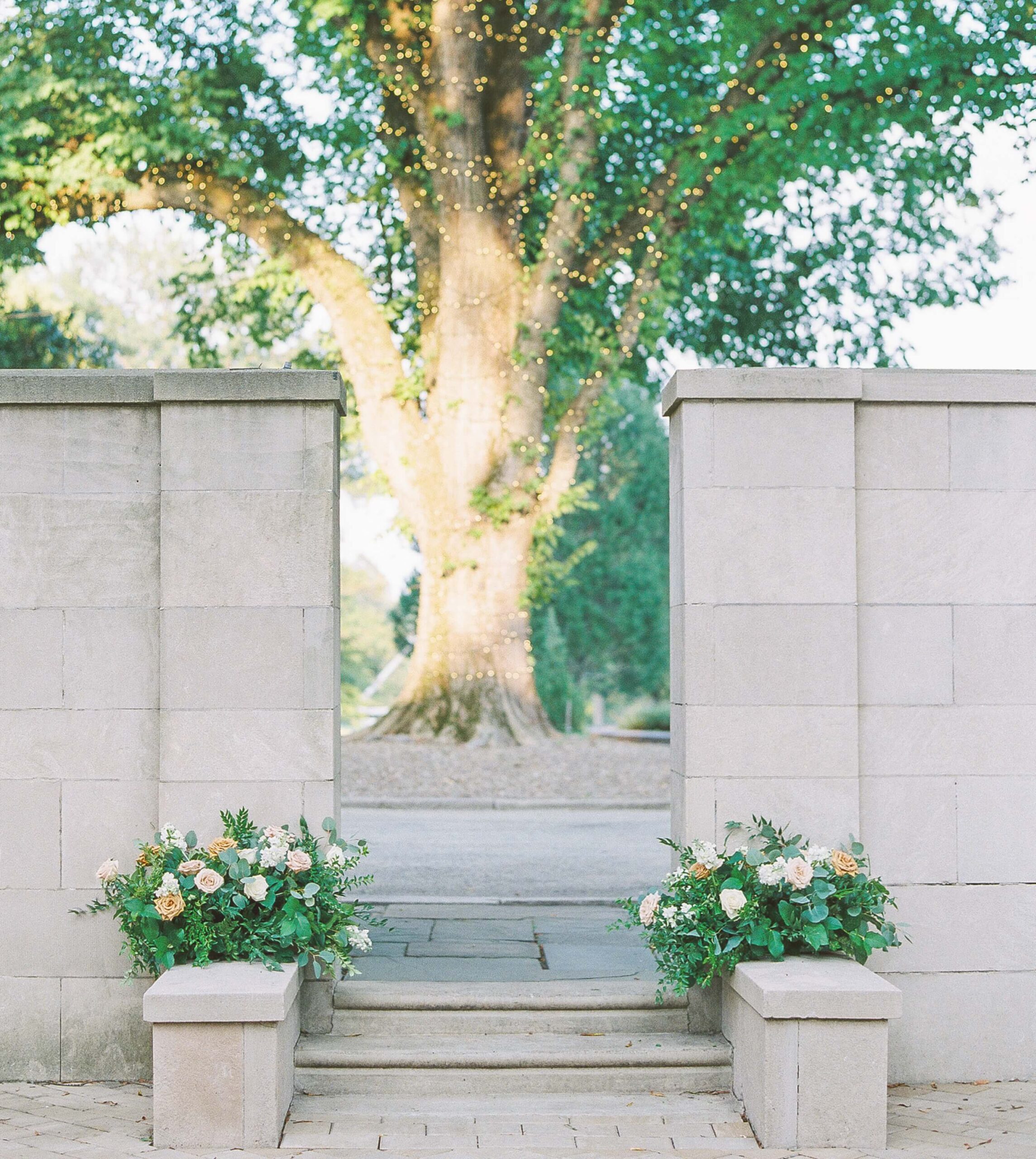 Stone entrance with two flower arrangements, leading to a large tree adorned with string lights.