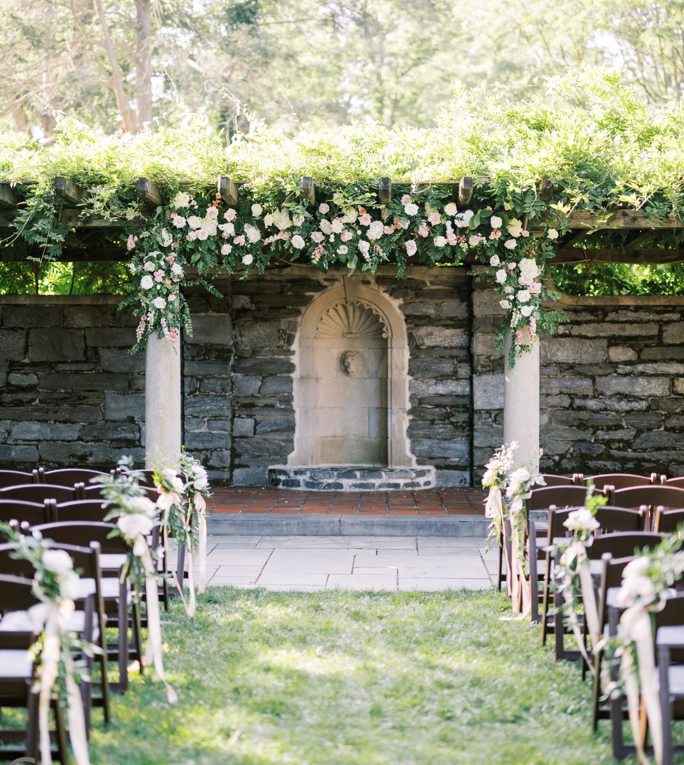 Outdoor wedding ceremony setup with rows of chairs facing a stone archway adorned with green and white floral arrangements.