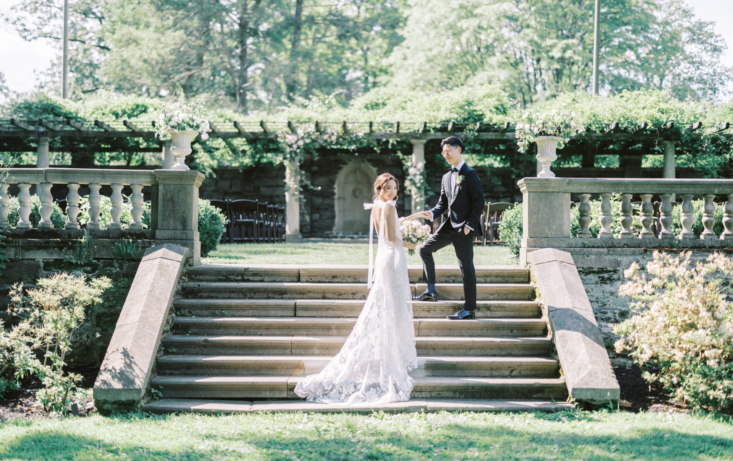 A bride in a white gown and a groom in a black suit stand on outdoor stone steps surrounded by greenery, holding hands.