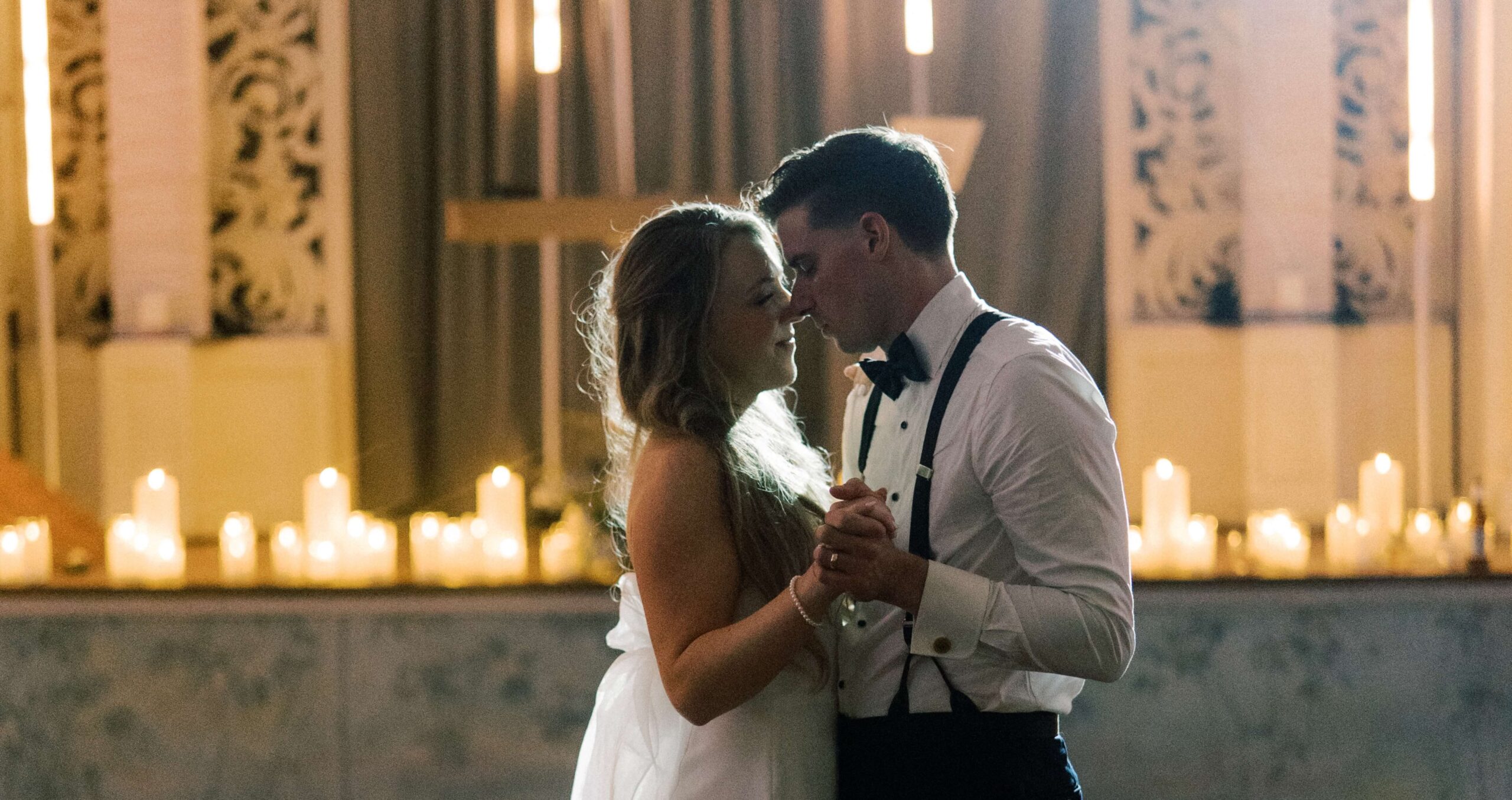 A couple dressed in formal wedding attire dances closely together in a softly lit room with candles in the background.