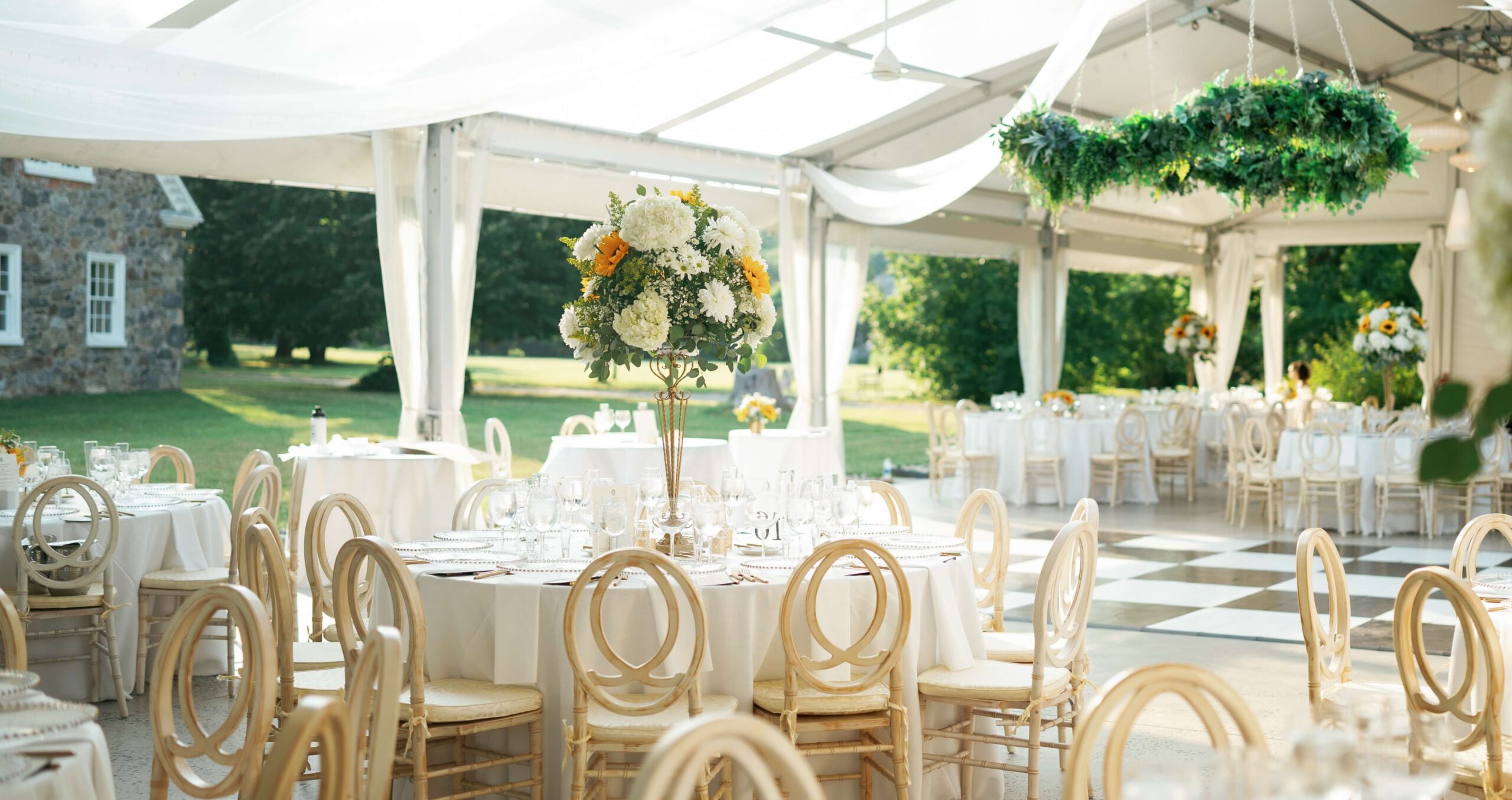 A decorated outdoor event space with round tables, floral centerpieces, beige chairs, and a checkered dance floor under a transparent tent.