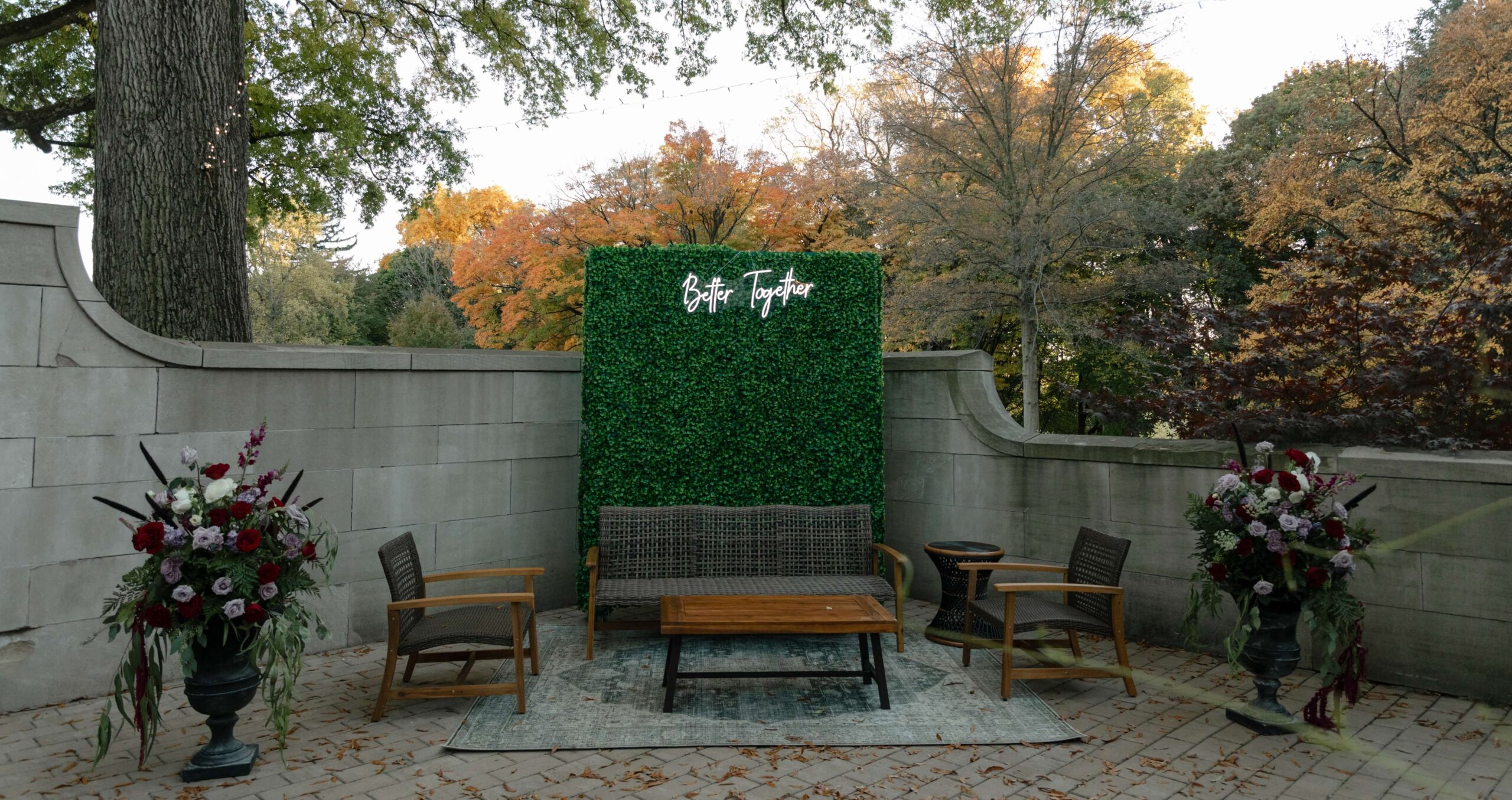 Outdoor seating area with wooden chairs, a loveseat, coffee table, potted flower arrangements, and a green hedge backdrop with a white neon sign reading 