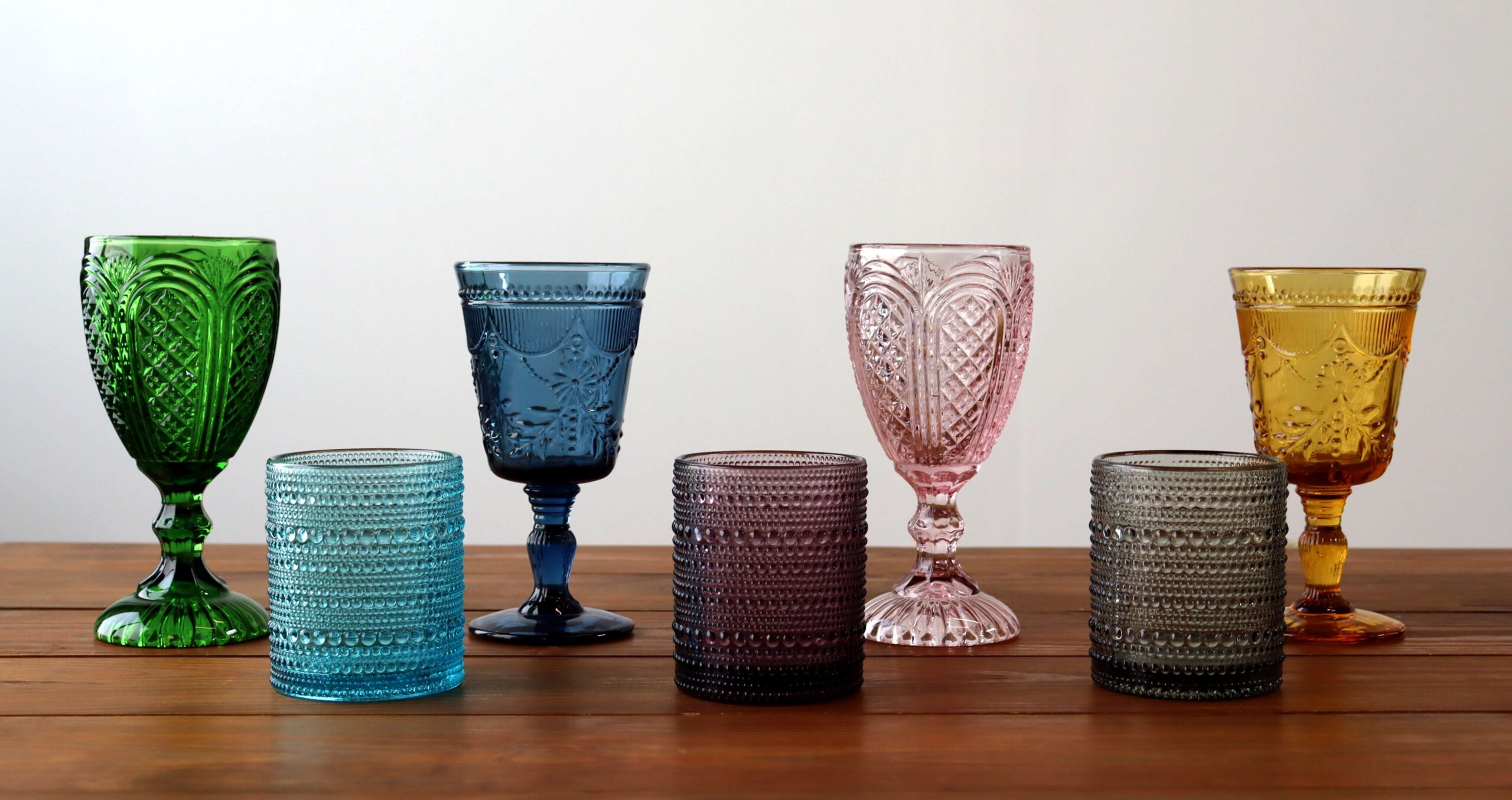 Six colored glasses, three goblets and three tumblers, are arranged in a row on a wooden table against a plain light background.