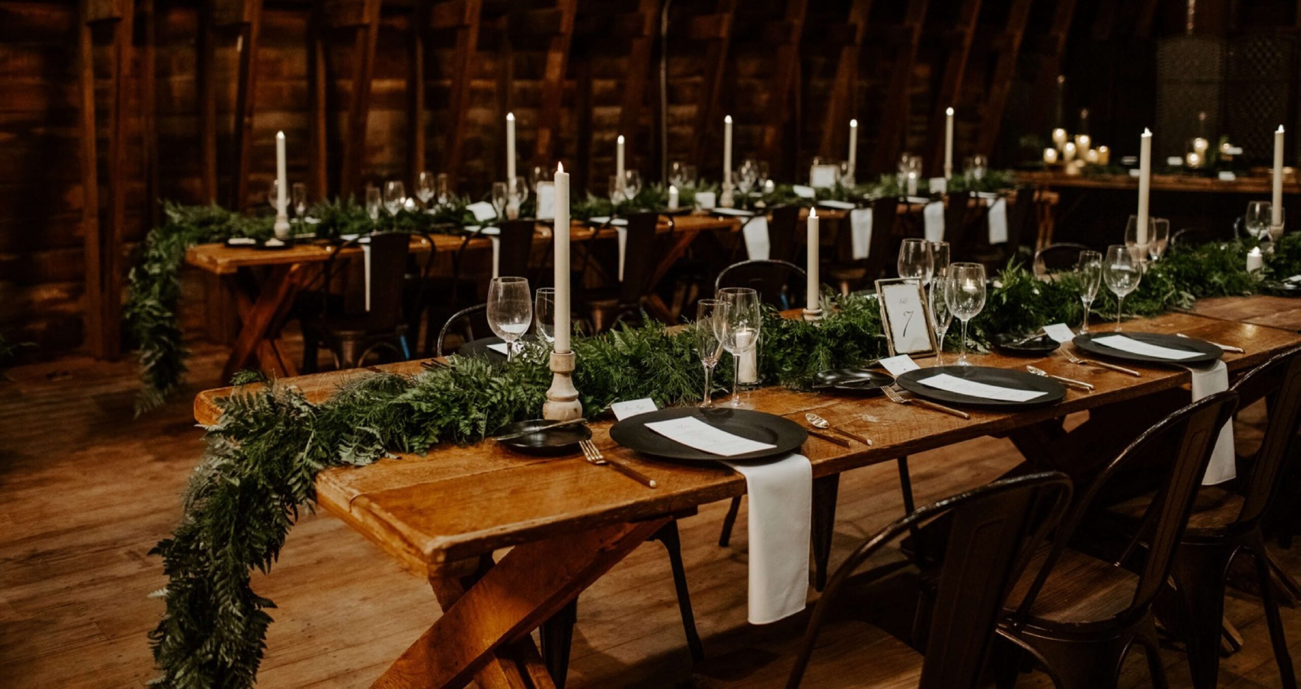 Long wooden tables set for a formal dinner with white plates, black napkins, glassware, and greenery garlands in a rustic wooden room with candles.