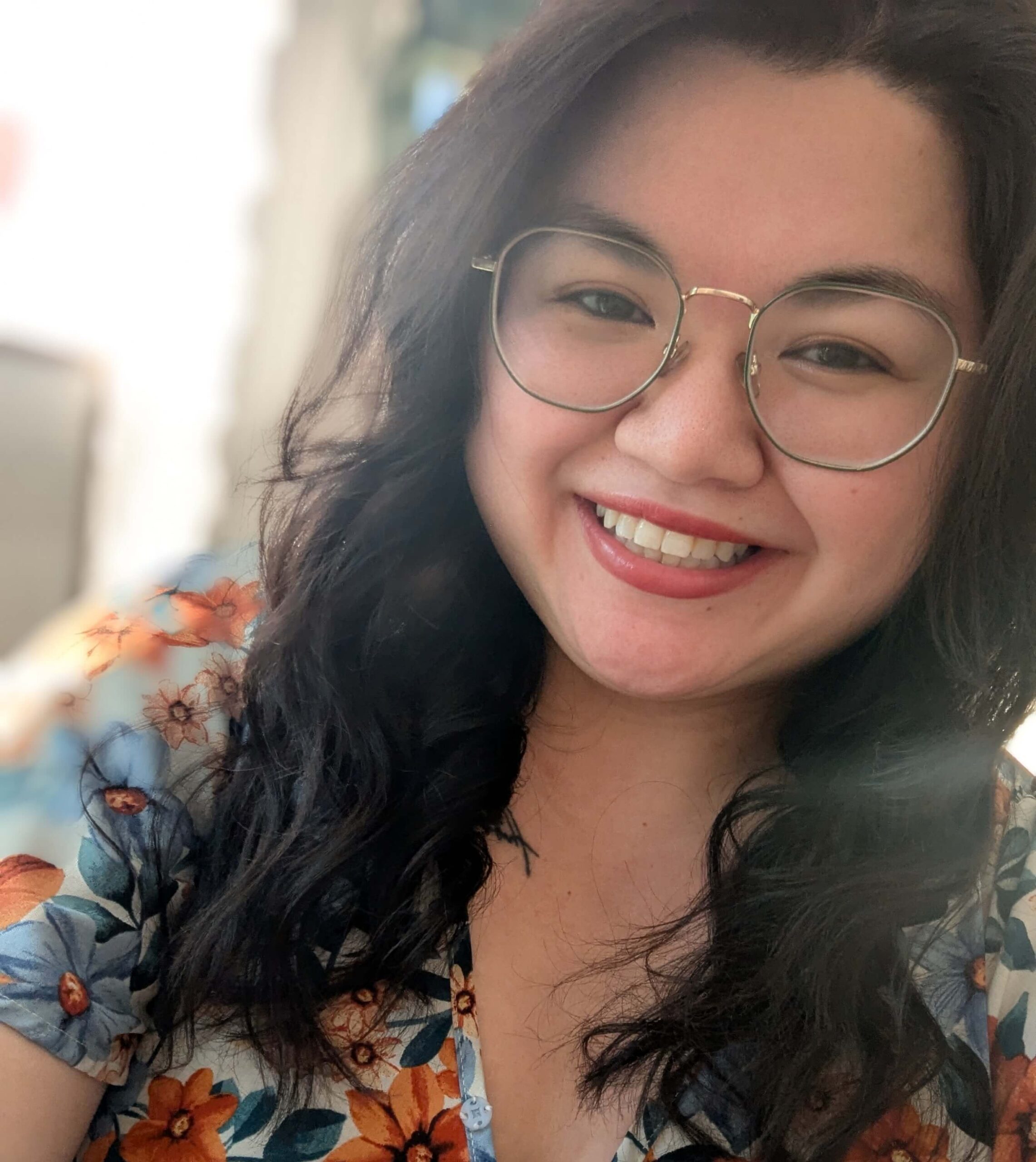 A woman with long dark hair, wearing glasses and a floral top, smiles at the camera in a well-lit indoor setting.