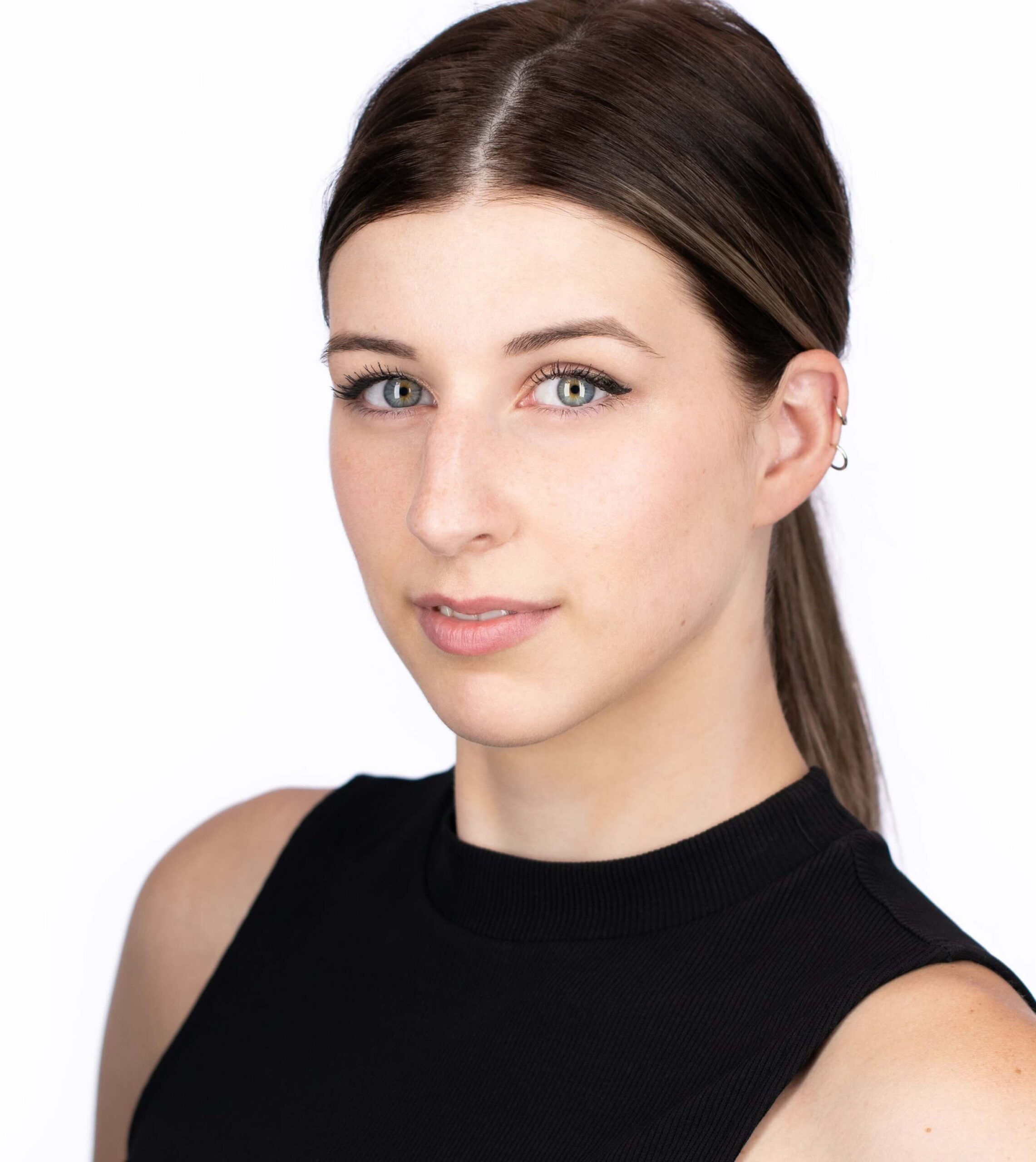 A woman with straight brown hair in a low ponytail, wearing a sleeveless black top, looks at the camera against a white background.