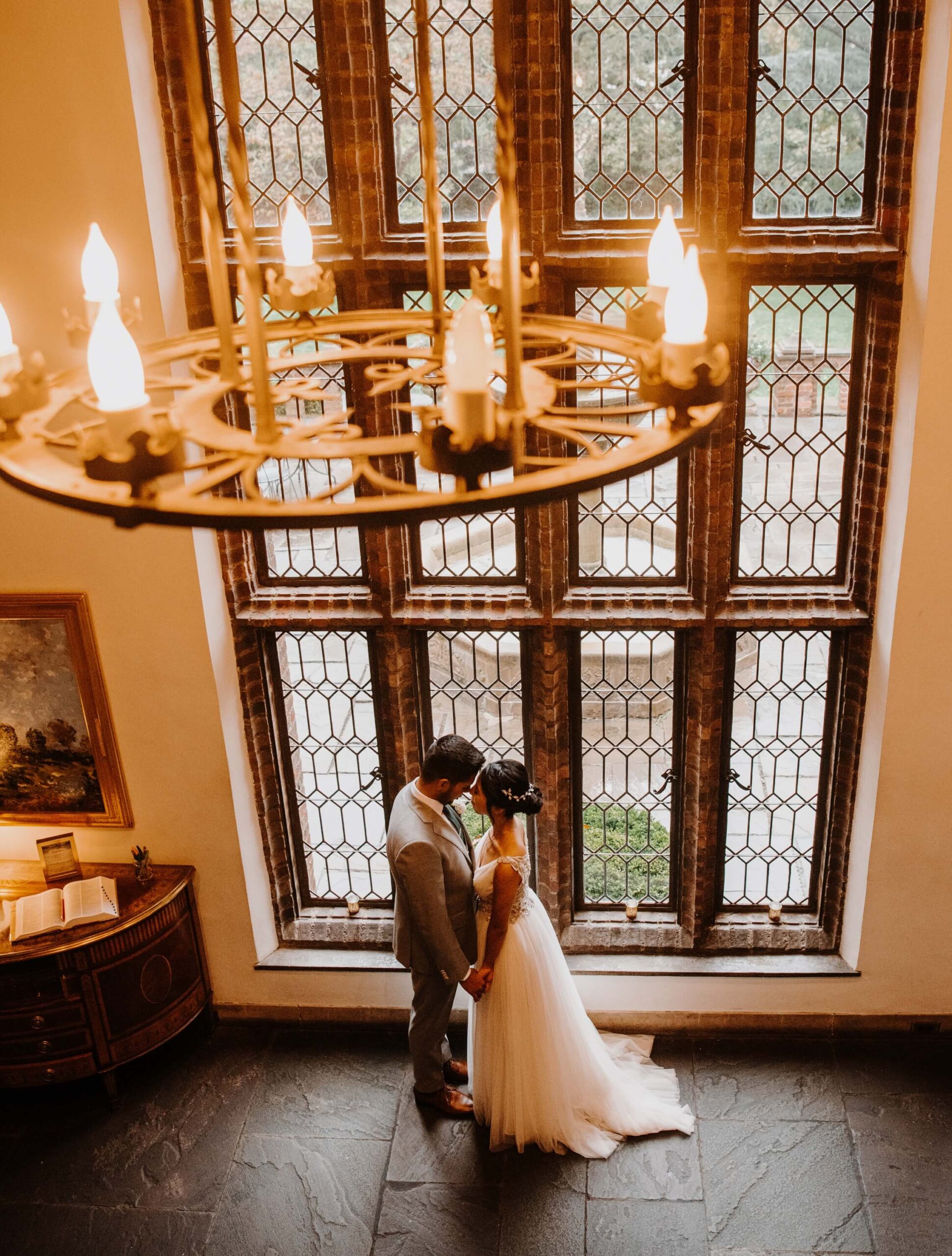 A bride and groom stand facing each other in front of tall, ornate windows, with a large chandelier hanging above them in a warmly lit room.