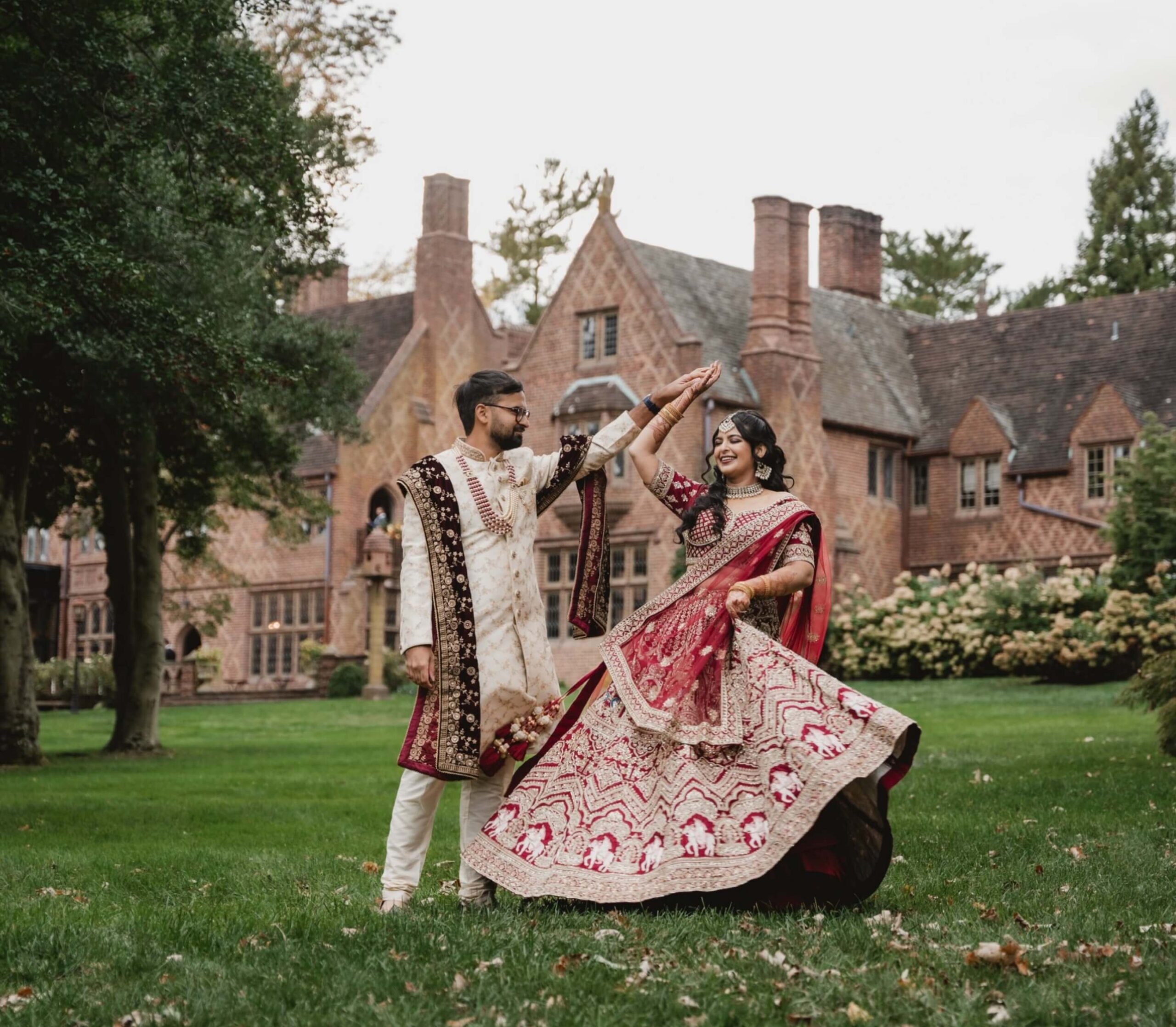 A couple in traditional Indian attire dance on a lawn in front of a large brick mansion, surrounded by trees and greenery.