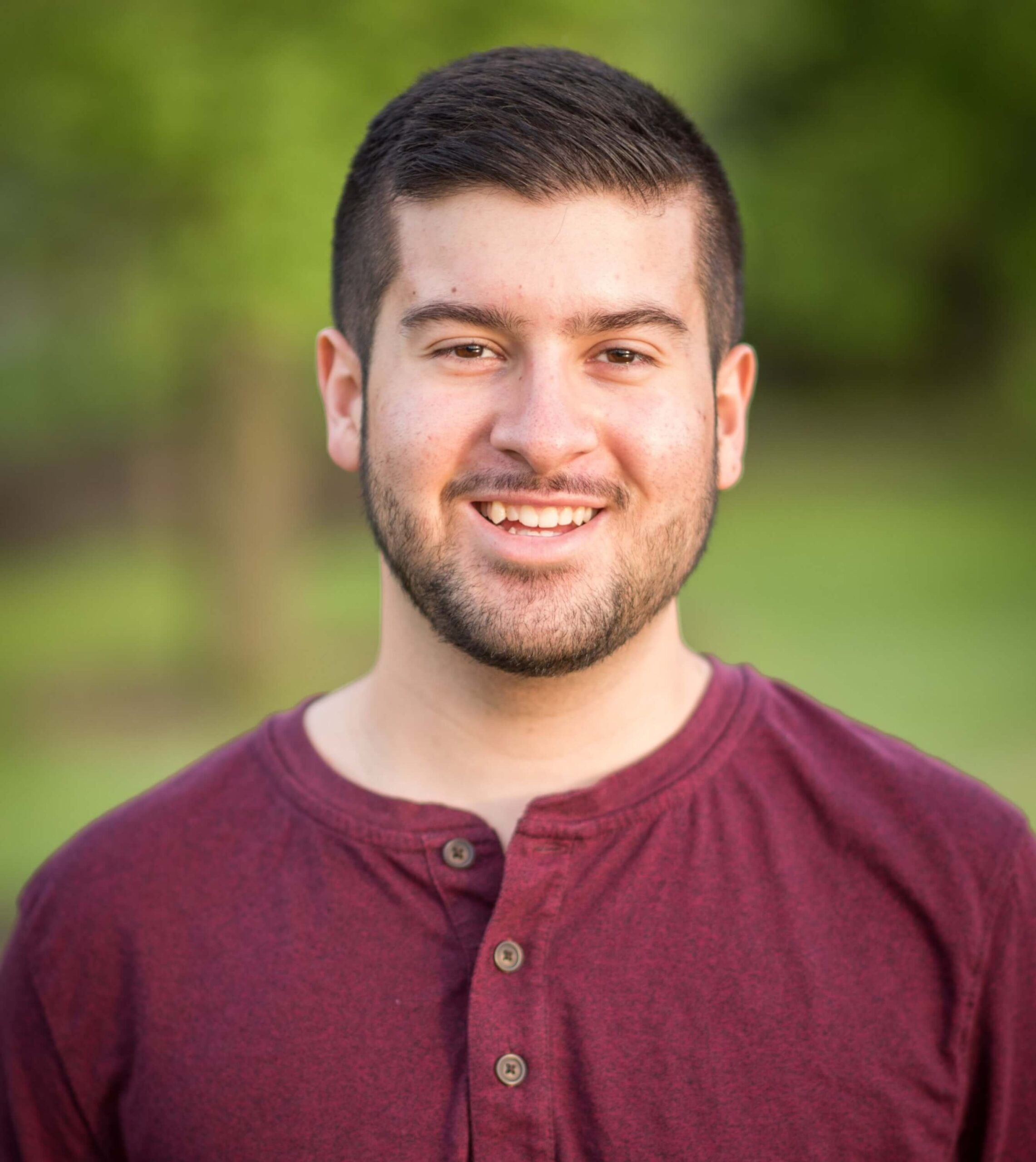 A young man with short dark hair and a trimmed beard smiles at the camera, wearing a maroon henley shirt. The background is blurred greenery.