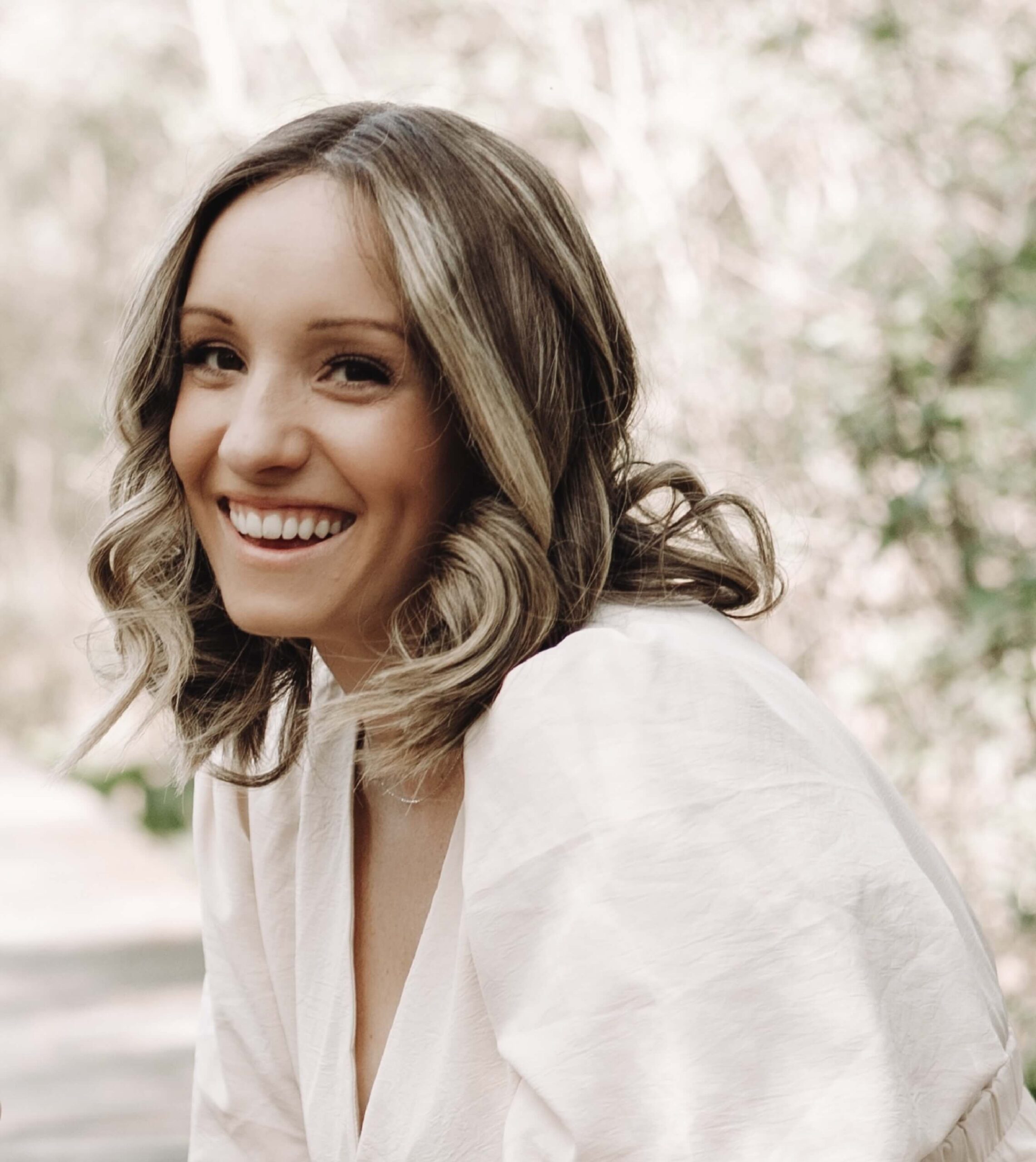 A woman with shoulder-length wavy hair and a white blouse smiles outdoors with trees in the background.