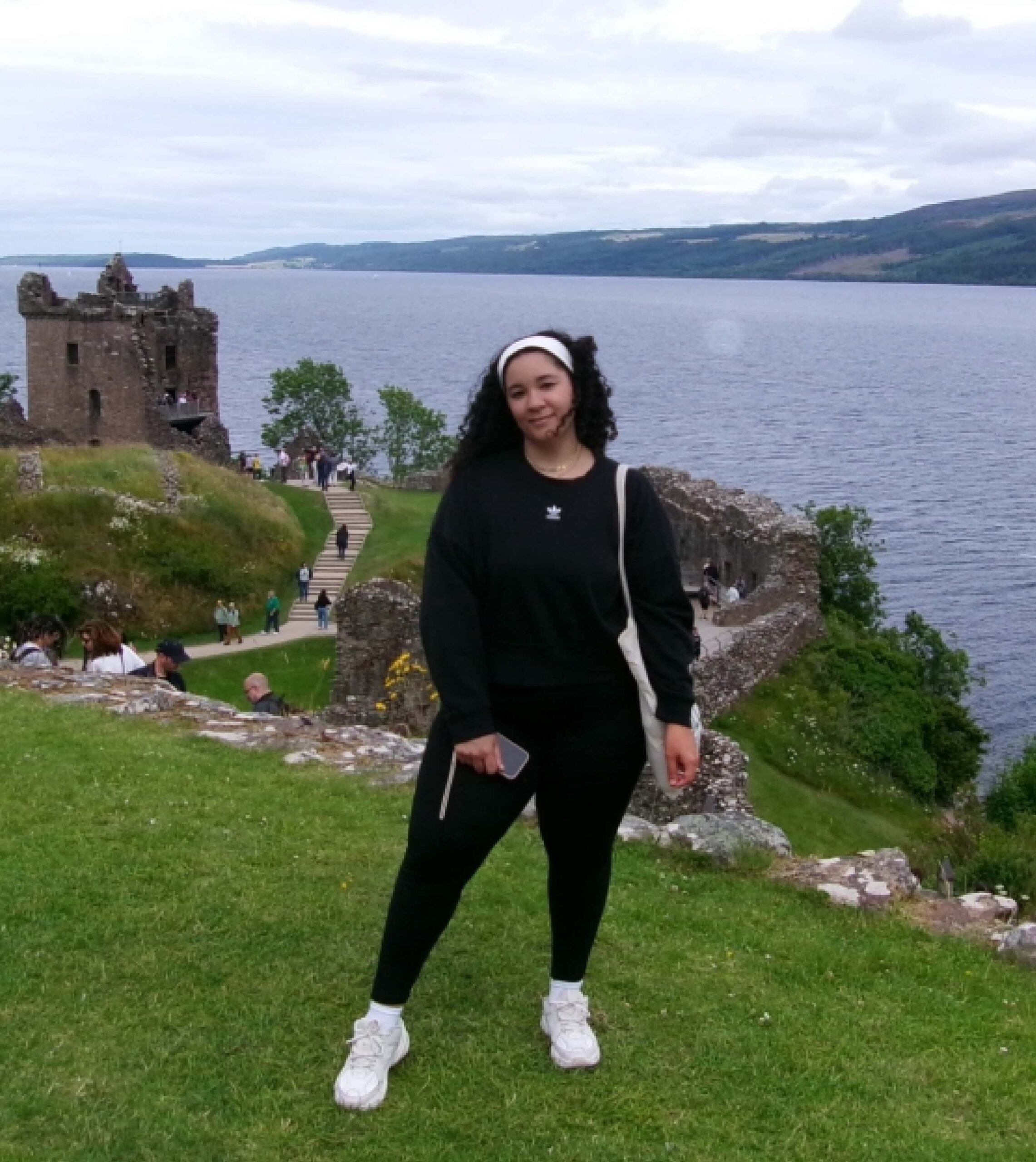A person stands on a grassy area in front of castle ruins by a large lake, with hills visible in the background under a cloudy sky.