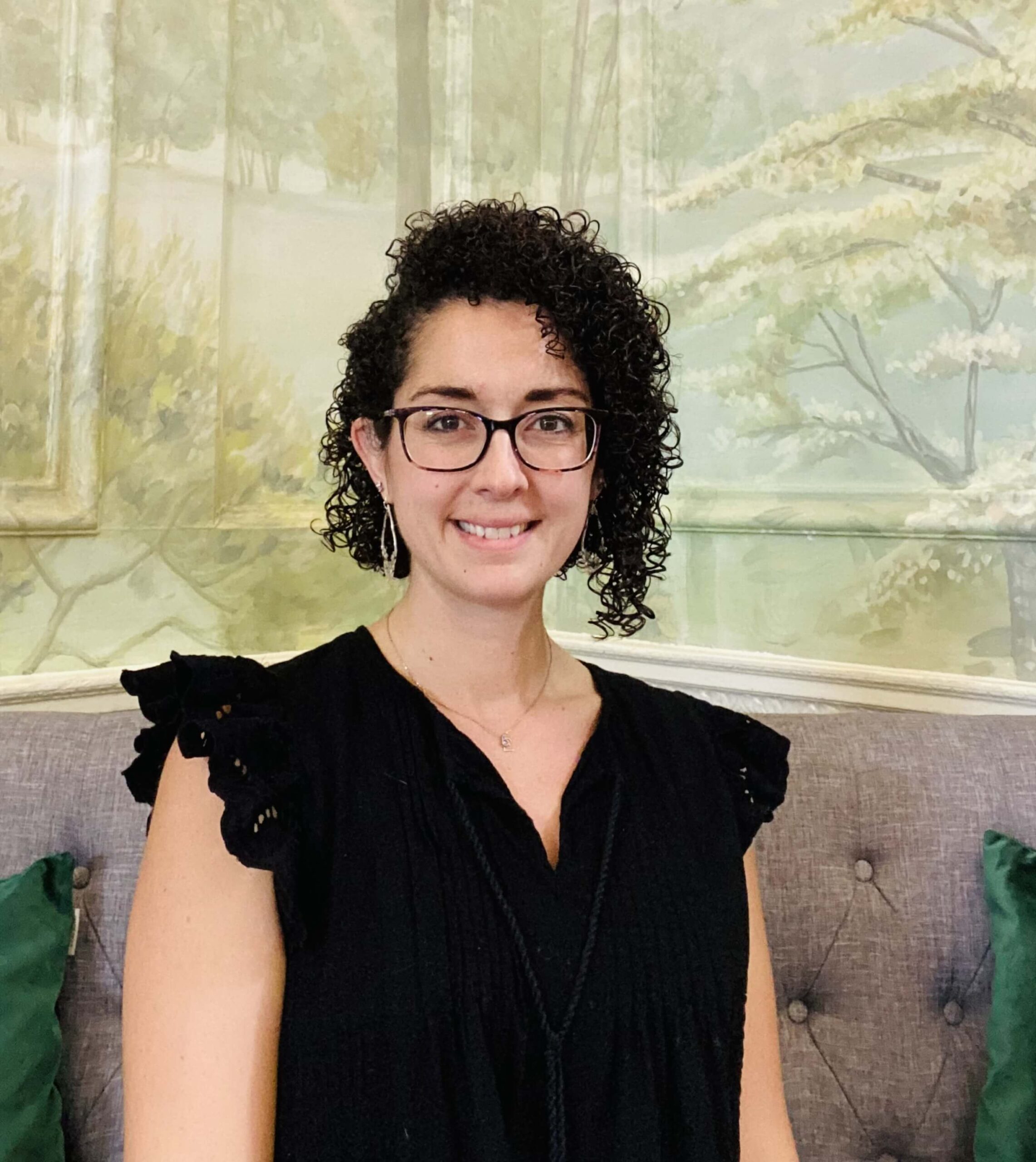 A woman with curly hair and glasses, wearing a black sleeveless top, sits on a gray tufted couch in front of a nature-themed mural.