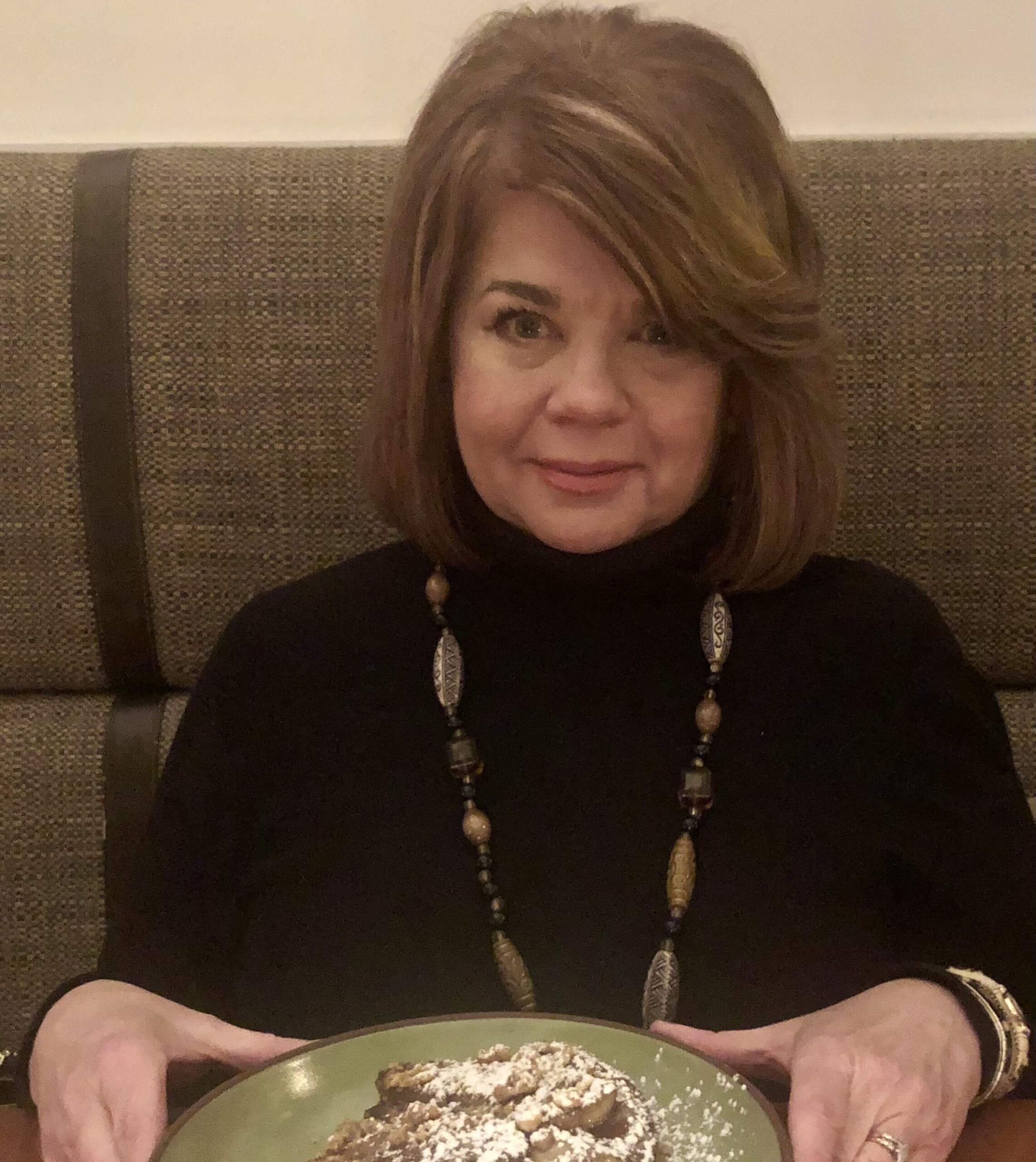 A woman with shoulder-length brown hair and a black top sits at a restaurant table, holding a plate of food topped with powdered sugar.