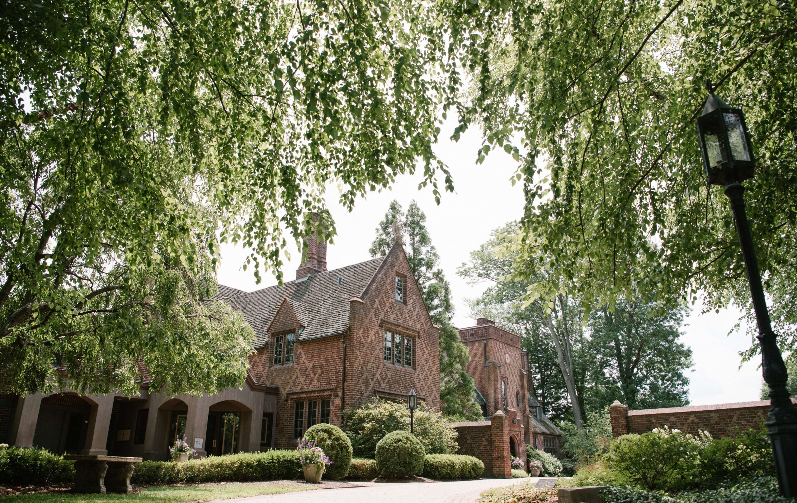 A large brick house with tall windows and a landscaped garden, framed by overhanging tree branches on a sunny day.