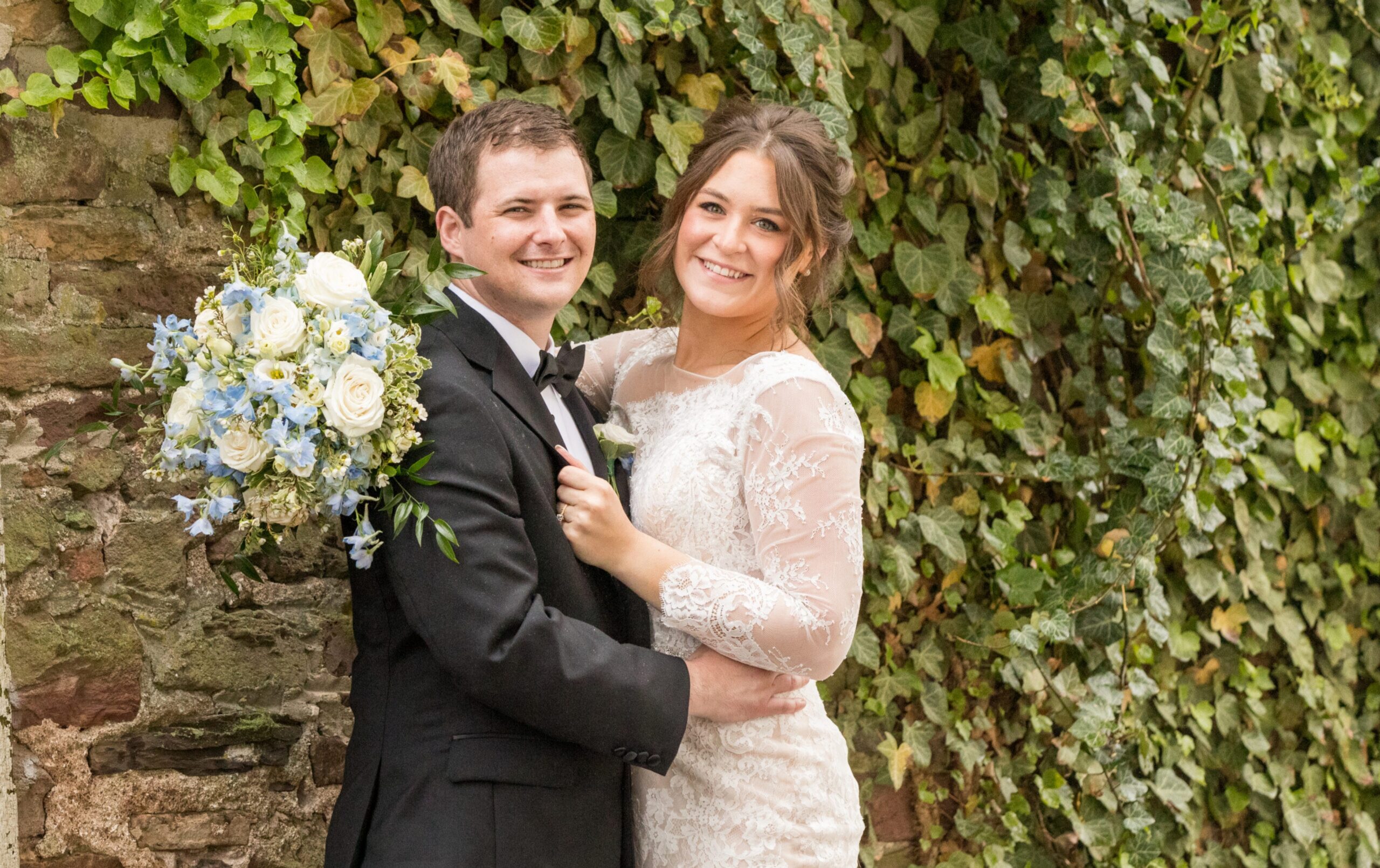 A smiling couple in formal wedding attire pose in front of an ivy-covered stone wall; the bride holds a bouquet of white and blue flowers.