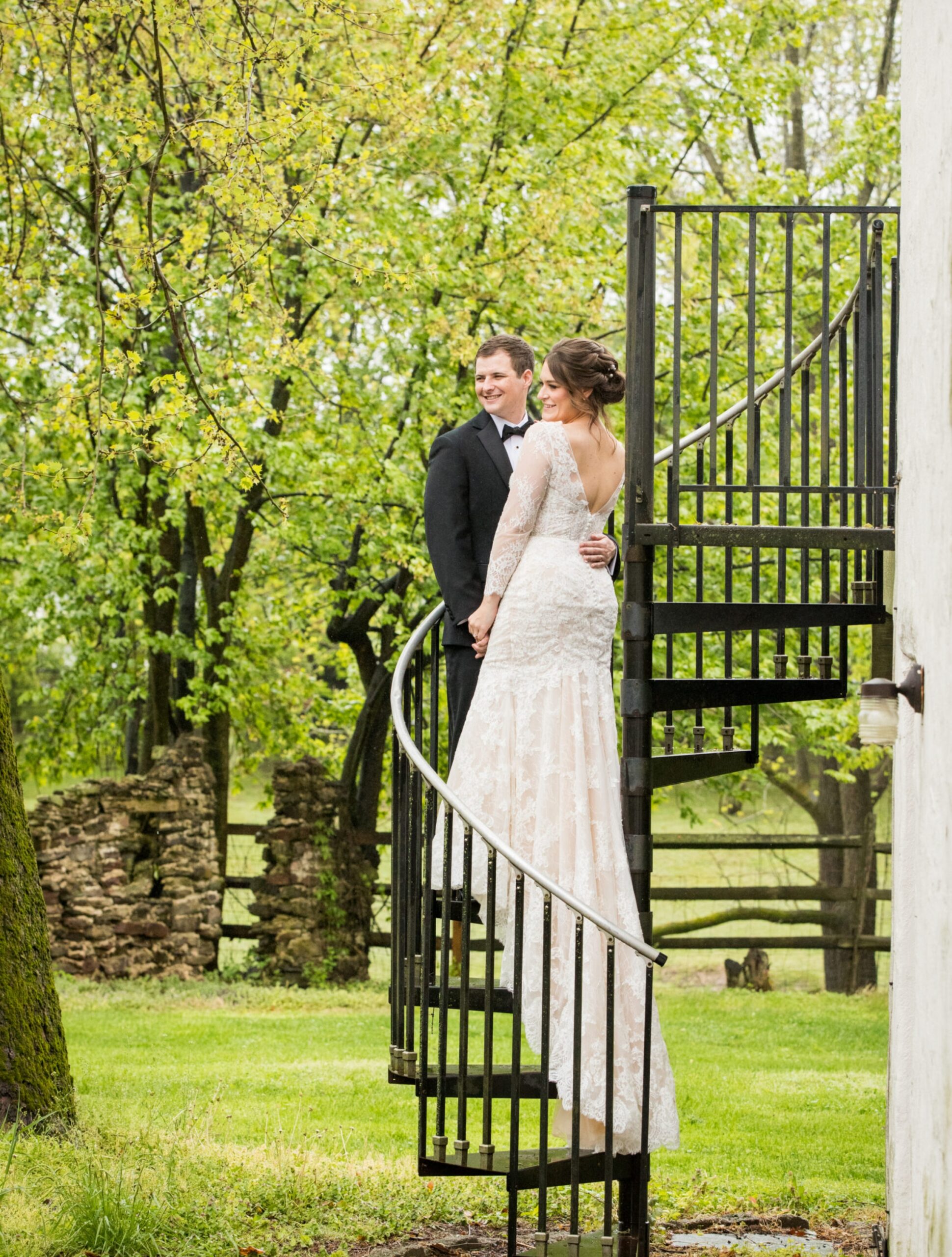 A bride and groom stand on a black spiral staircase outdoors, with green trees and a stone fence in the background.