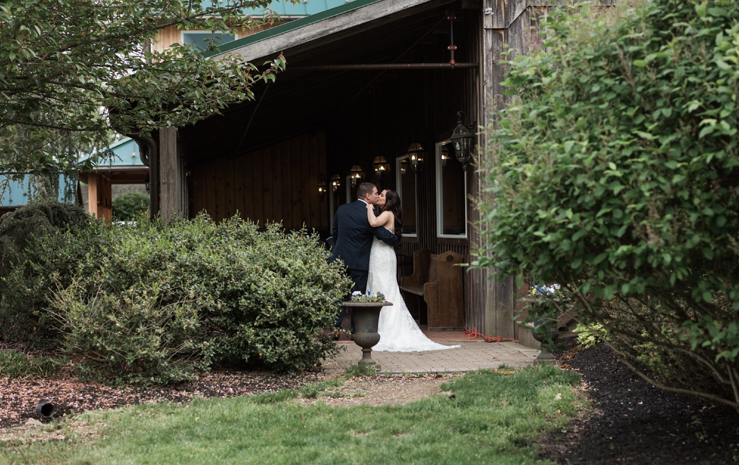 A bride and groom in wedding attire share a kiss on a garden path outside a rustic wooden building, surrounded by greenery.