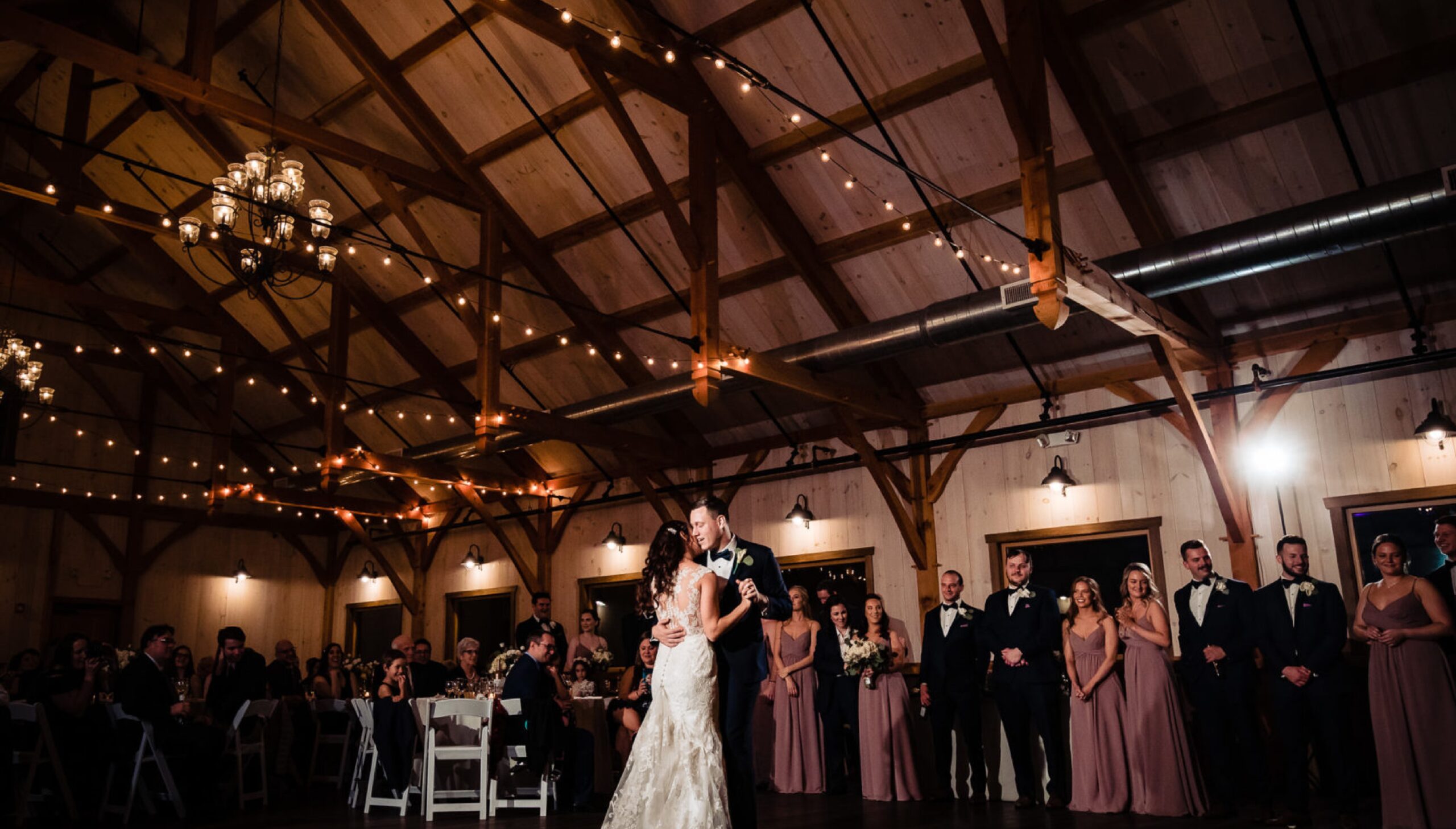 A bride and groom share their first dance in a rustic barn venue, surrounded by their wedding party and guests seated at tables under string lights.