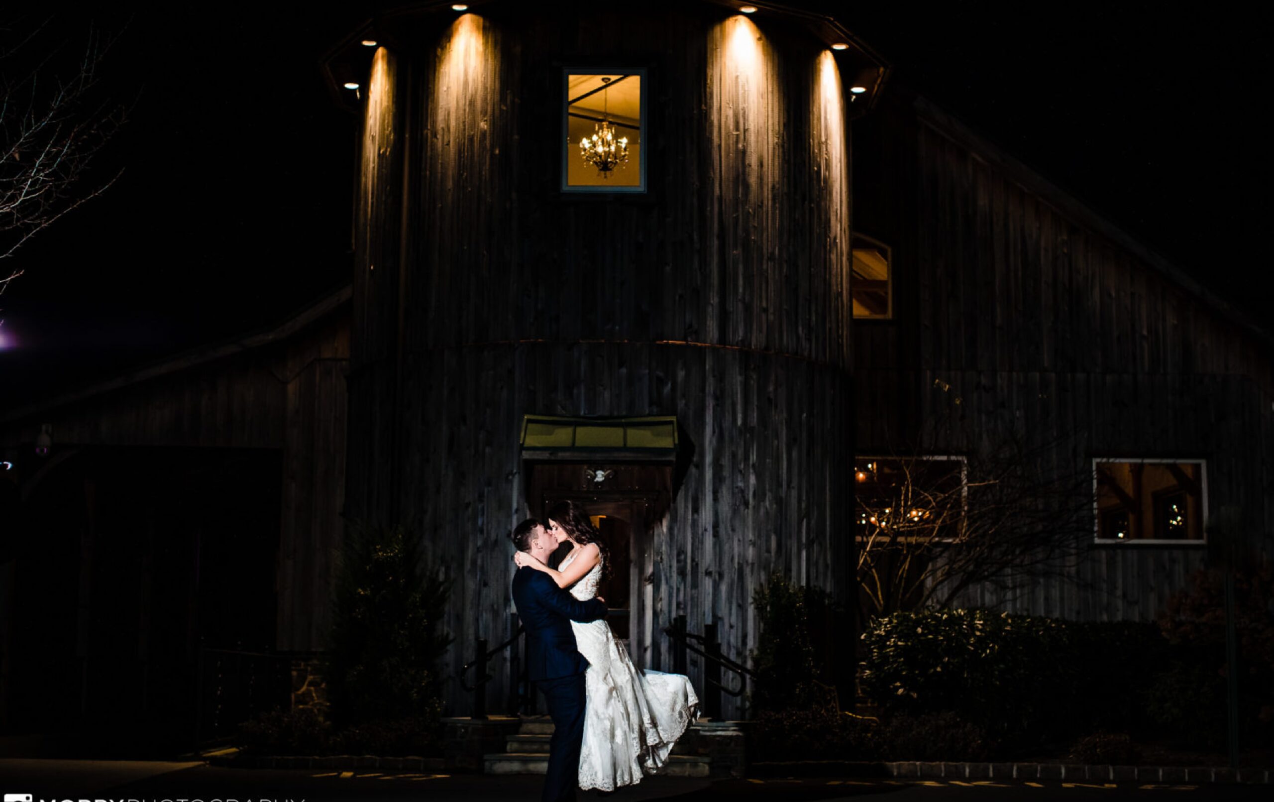 A groom lifts and kisses the bride at night in front of a wooden barn-style building illuminated by exterior lights.