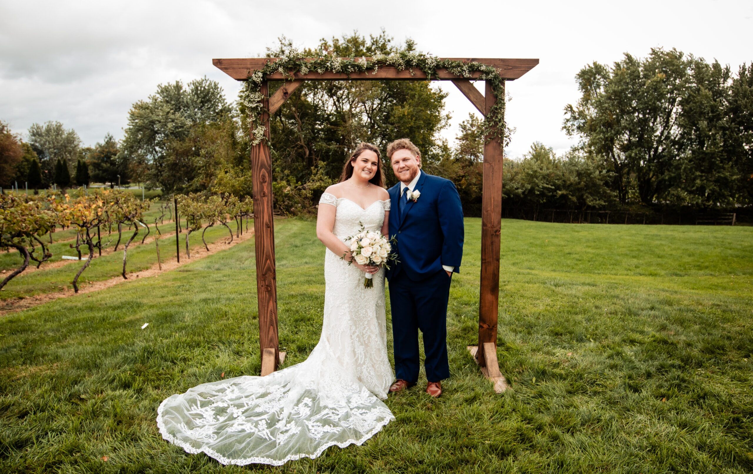 A bride in a lace wedding dress and a groom in a blue suit stand under a wooden arch on a grassy lawn, smiling at the camera.