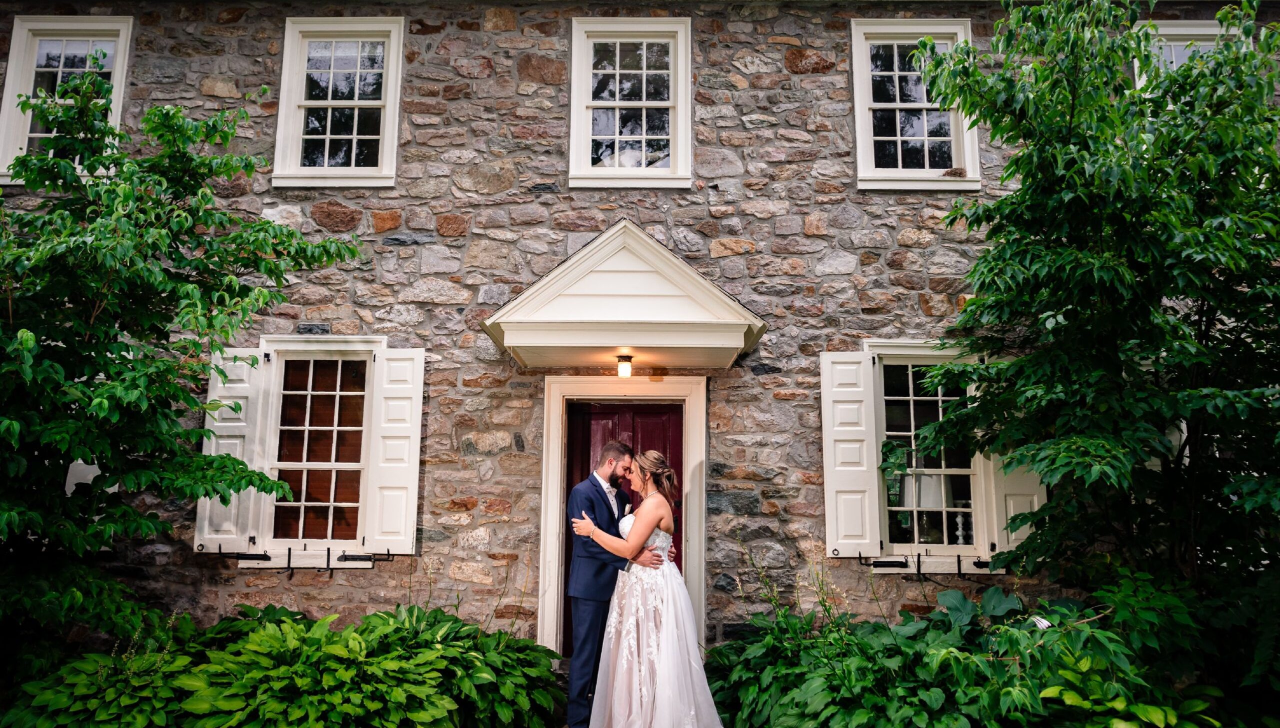 A couple dressed in wedding attire embrace and kiss in front of a stone house with white trim, surrounded by greenery.
