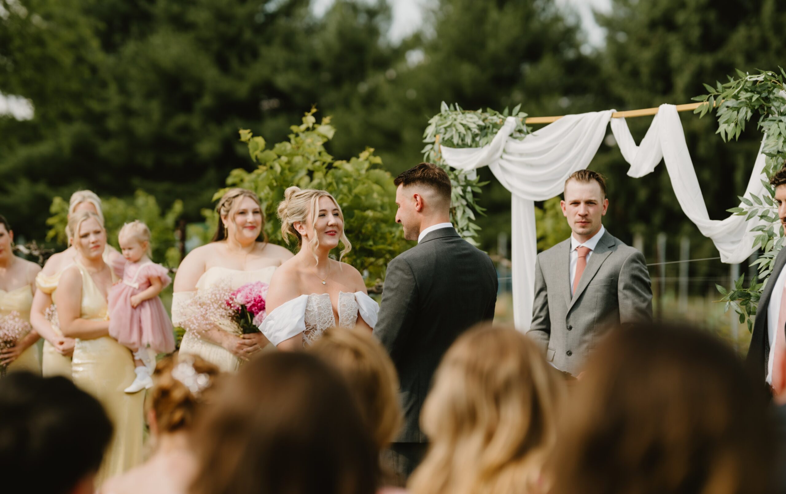 A bride and groom stand facing each other during an outdoor wedding ceremony, with bridesmaids, a groomsman, and guests present.