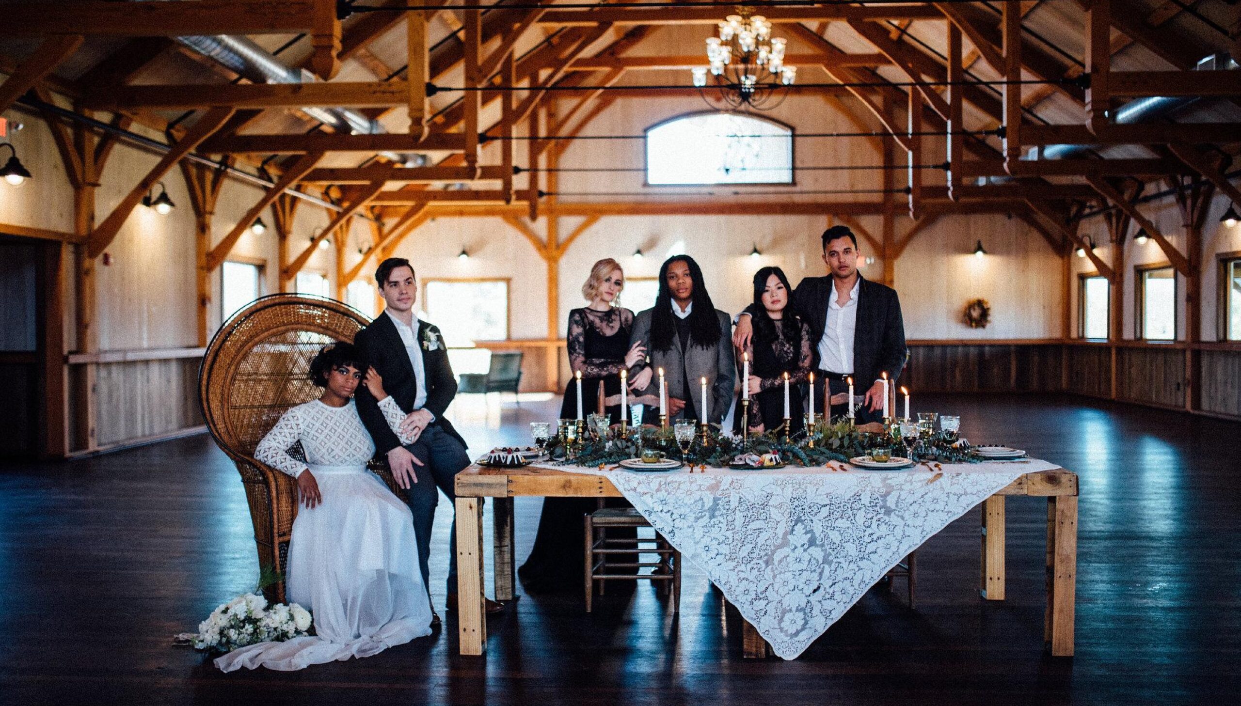 Six people in formal attire pose around a decorated dining table in a spacious wooden hall with exposed beams and chandeliers.