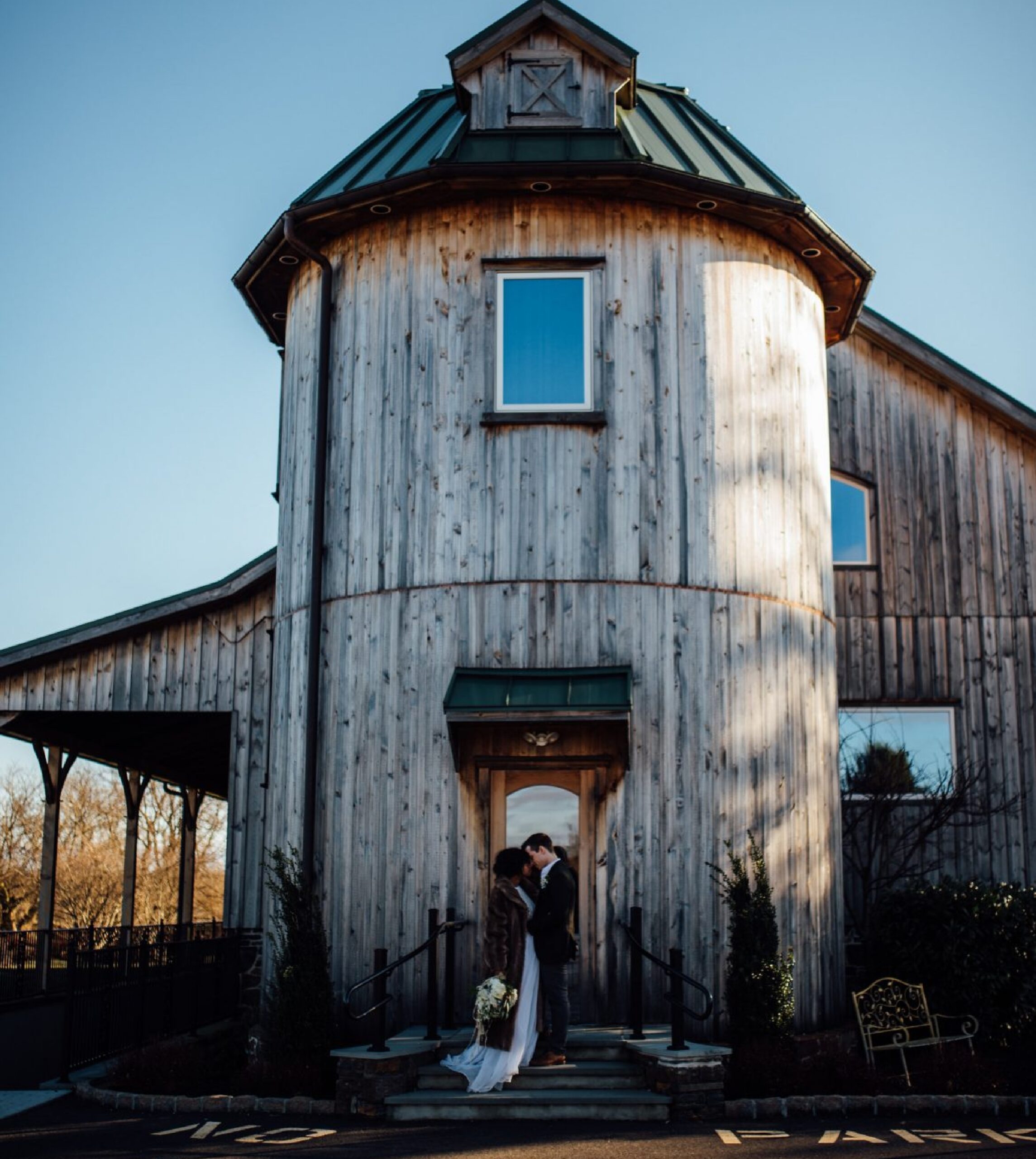 A couple in wedding attire stands embracing at the entrance of a rustic, cylindrical wooden building under a clear blue sky.