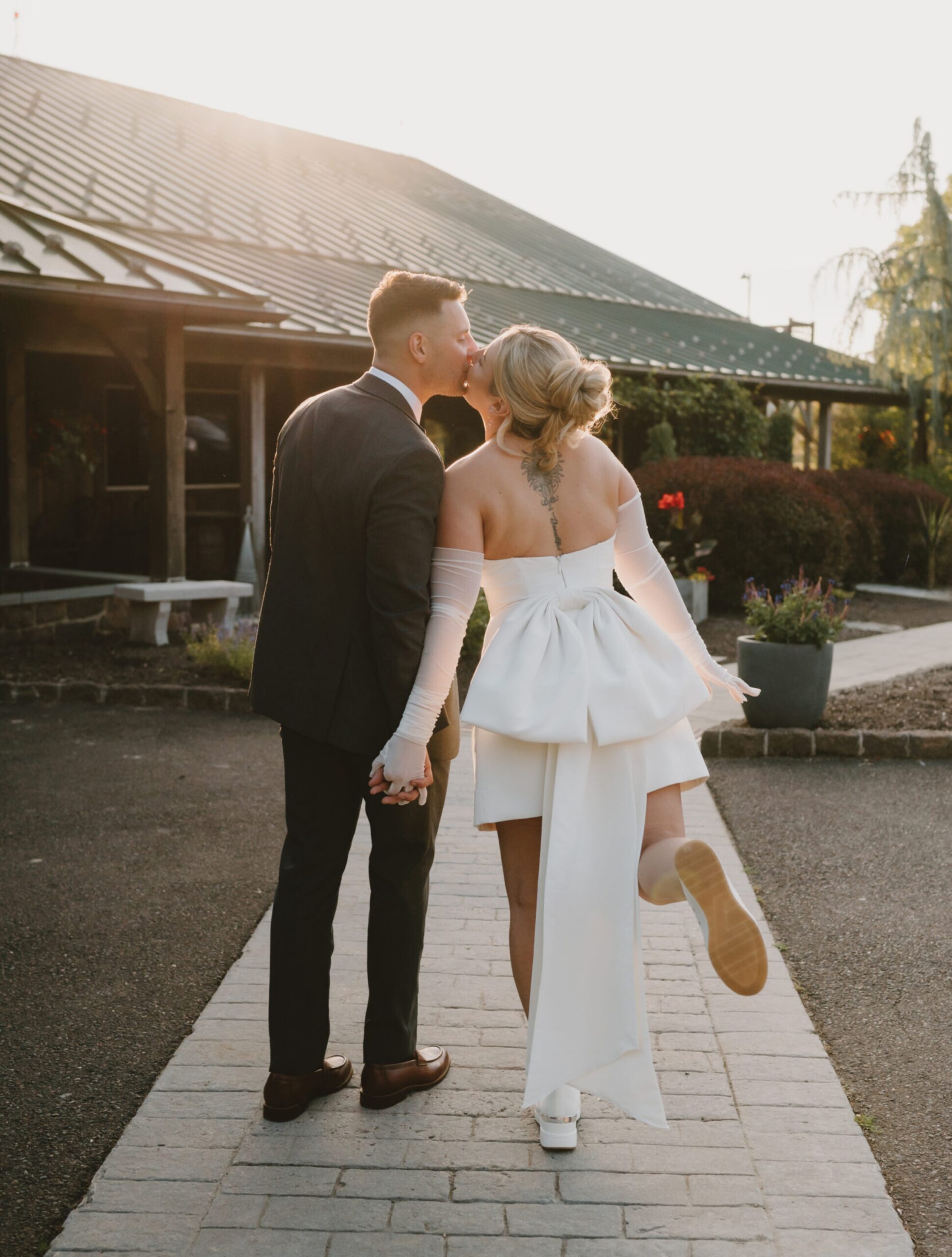 A couple dressed in wedding attire walks hand in hand on a paved path; the bride kicks up her heel as they share a kiss.