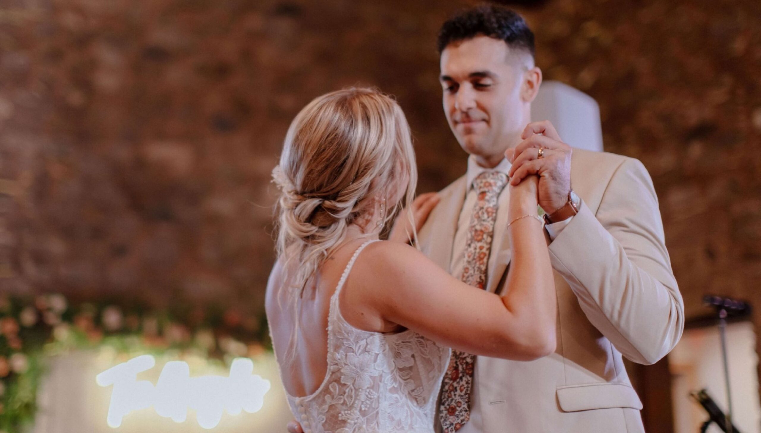 A bride and groom dance together, holding hands, with the groom wearing a beige suit and floral tie and the bride in a lace dress. A blurred sign and flowers are visible in the background.