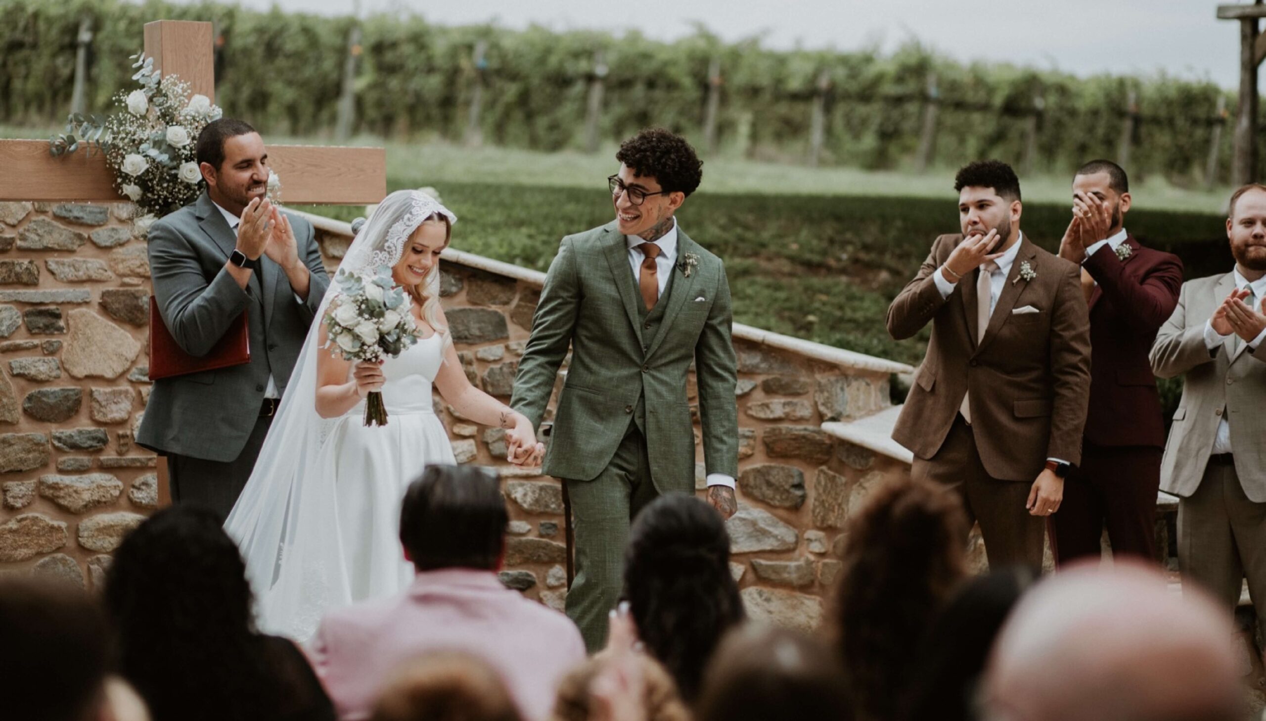 A bride and groom hold hands and smile at an outdoor wedding ceremony while guests and groomsmen applaud.