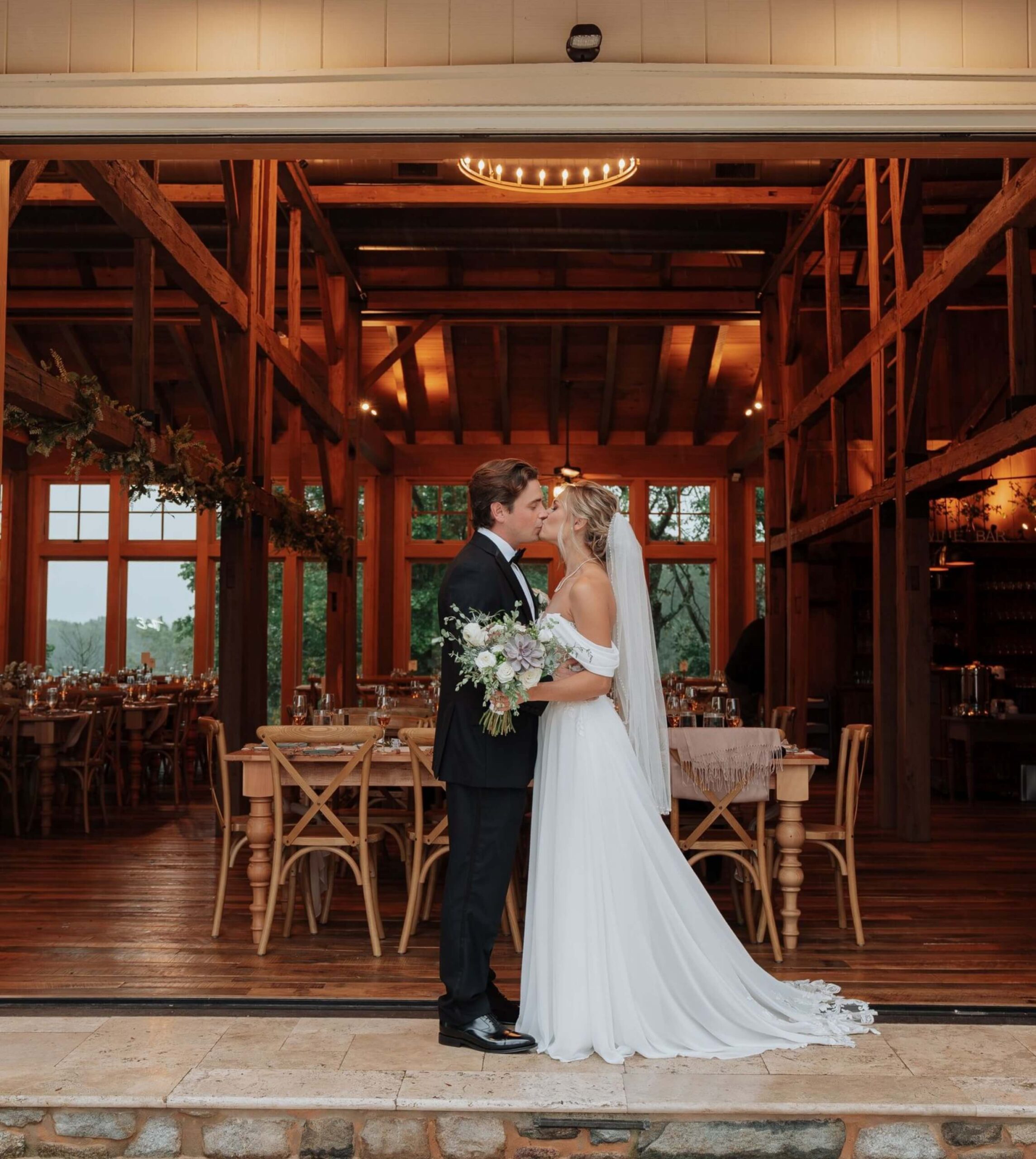 A bride and groom stand facing each other in front of a rustic wooden venue, holding a bouquet and dressed in wedding attire.