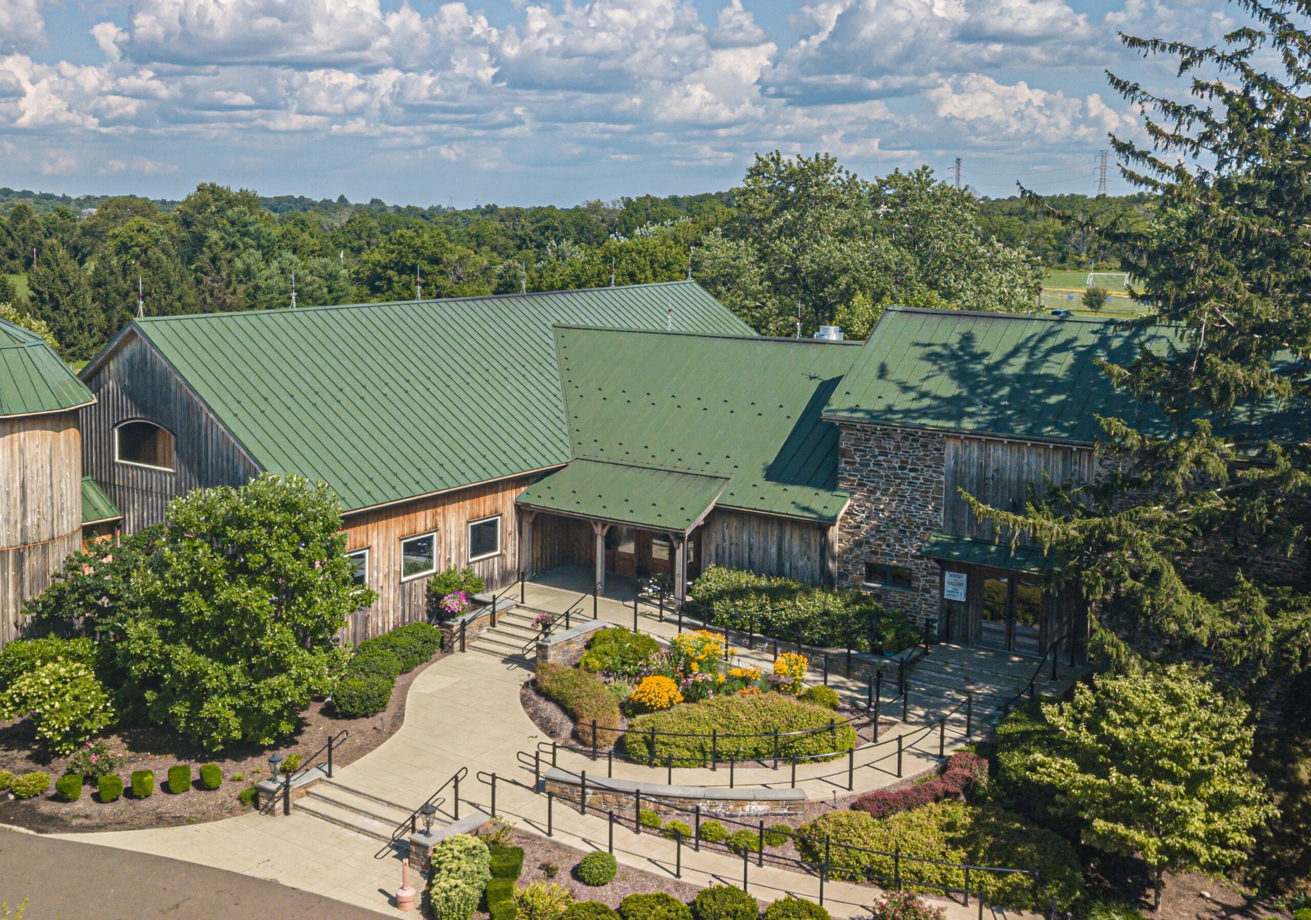 A large building with green metal roofs, stone and wood exterior, surrounded by landscaped shrubs and trees under a partly cloudy sky.