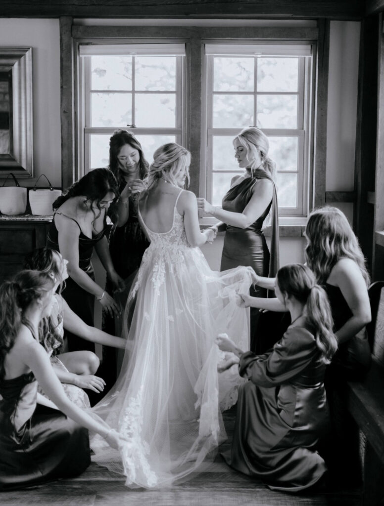 A bride stands in a room while bridesmaids in dark dresses help arrange her gown and veil. Natural light comes through the windows behind them. The image is in black and white.